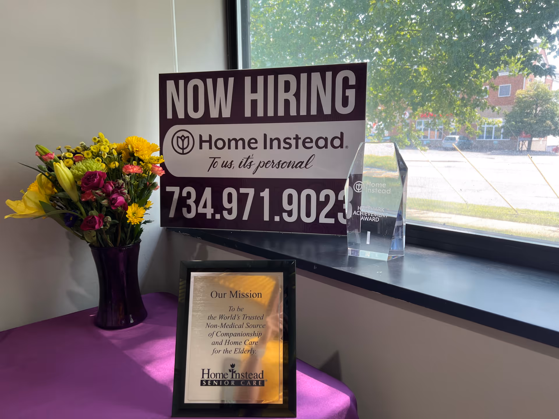 Indoor lobby corner with a Now Hiring sign, flowers, and a framed mission plaque.