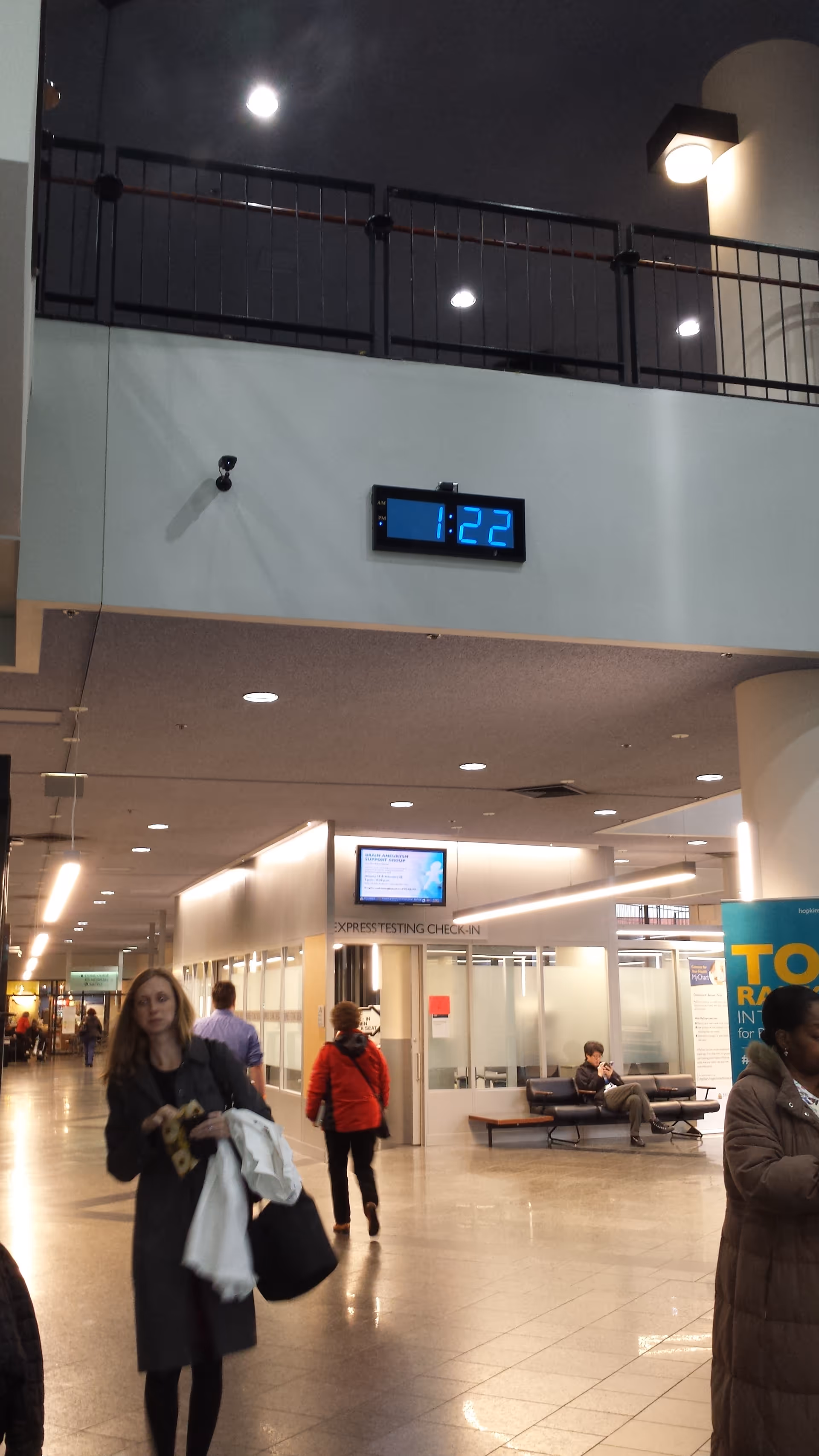 Interior hospital lobby with clock, check-in desks, seating, and people walking.