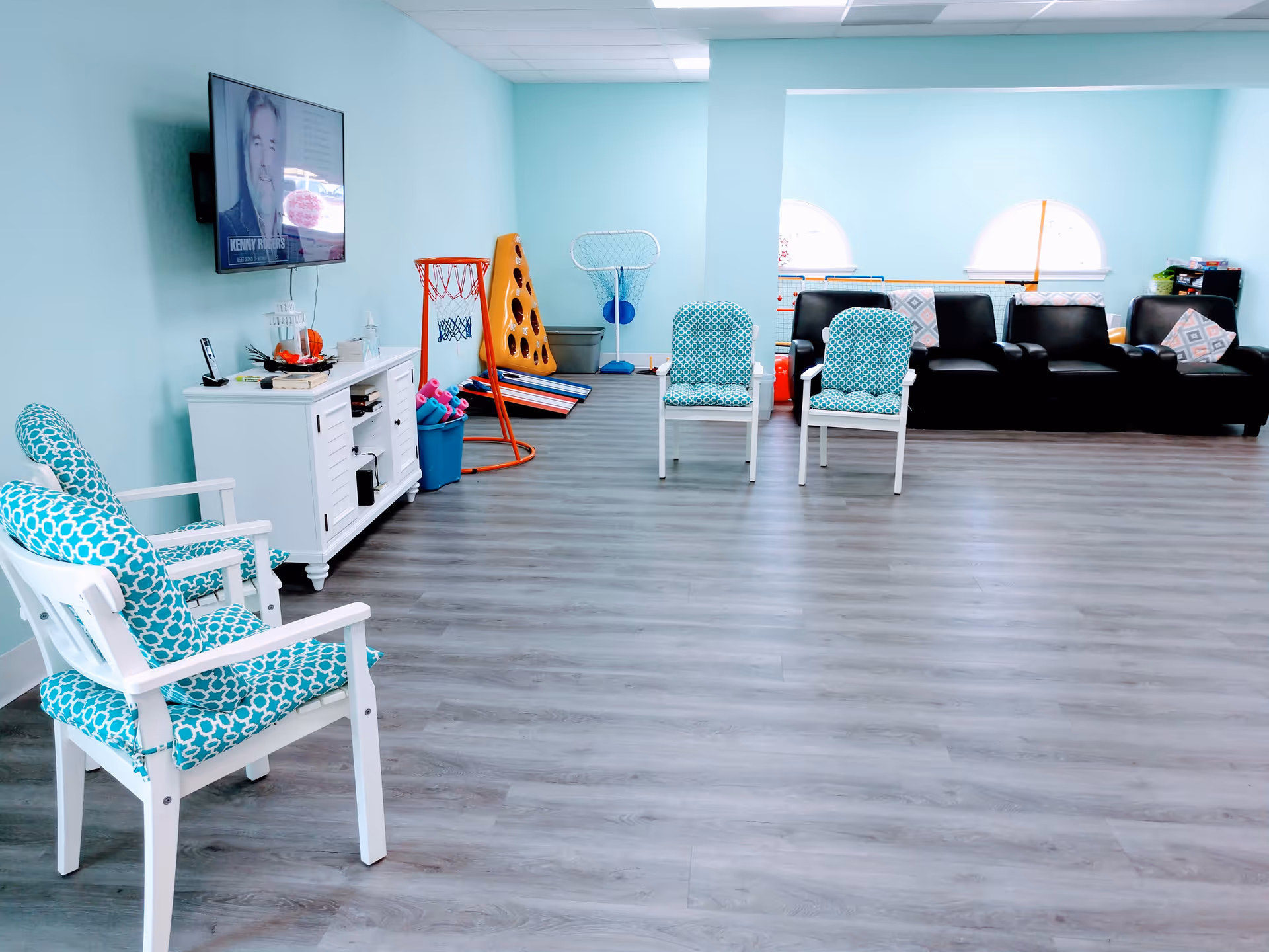 Bright activity lounge with turquoise chairs, a TV, and play equipment along the wall.