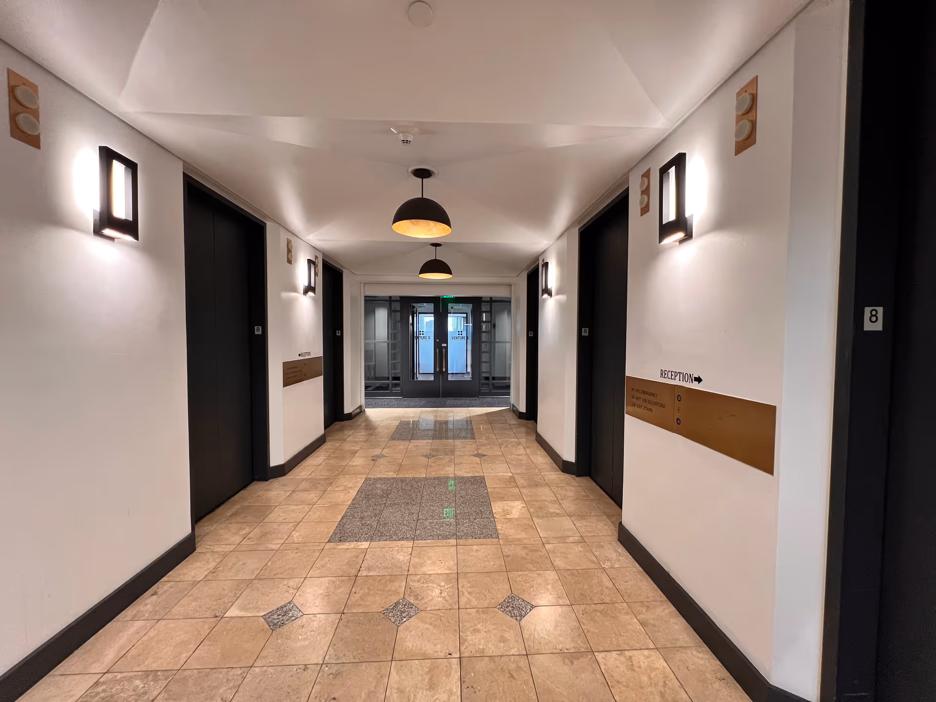 Bright hallway in a senior care facility with doors, tiled floor, and wall-mounted lights.