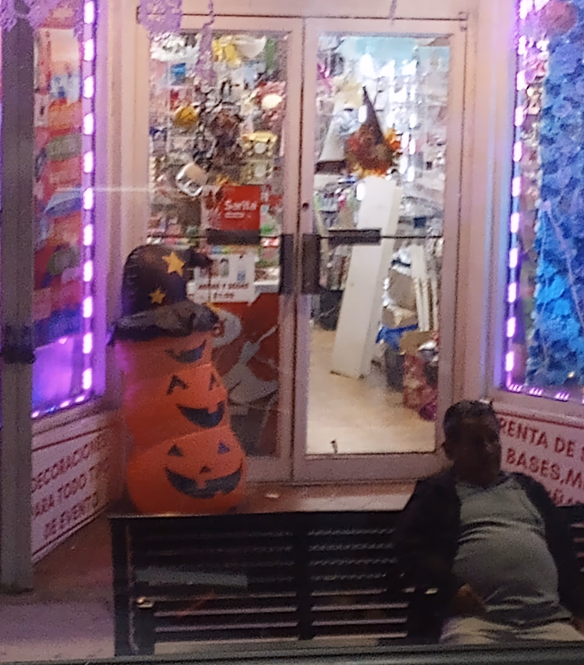 Glass storefront with purple-lit edges, Halloween pumpkin decorations, and a person sitting on a bench outside.