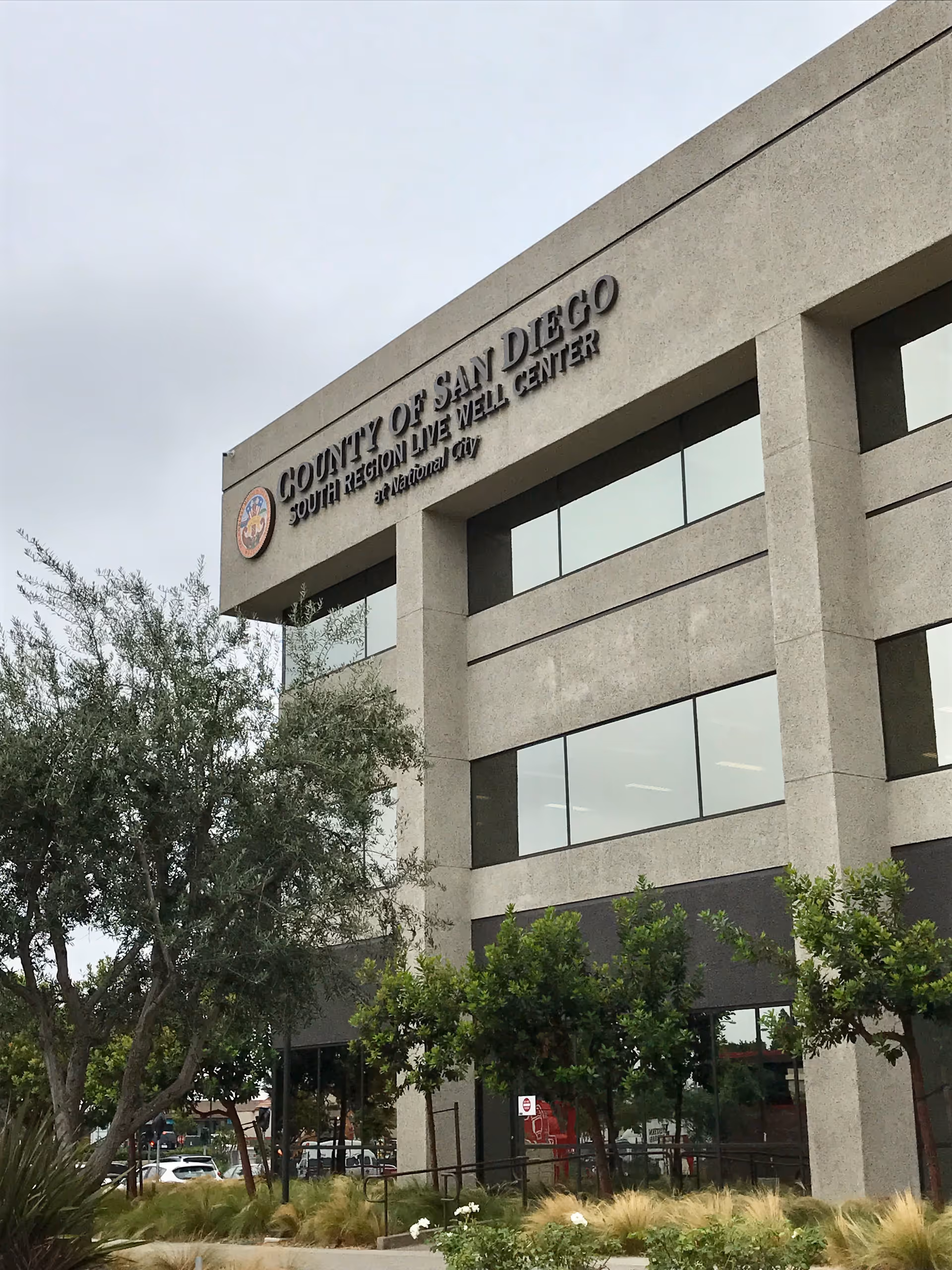 Front exterior of beige building with large windows and landscaping; sign reads County of San Diego.