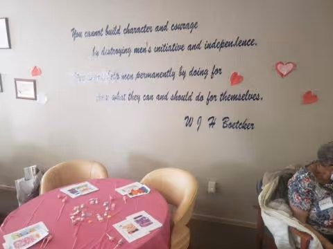 A round dining table with a pink tablecloth and chairs in a lounge, wall quote visible.