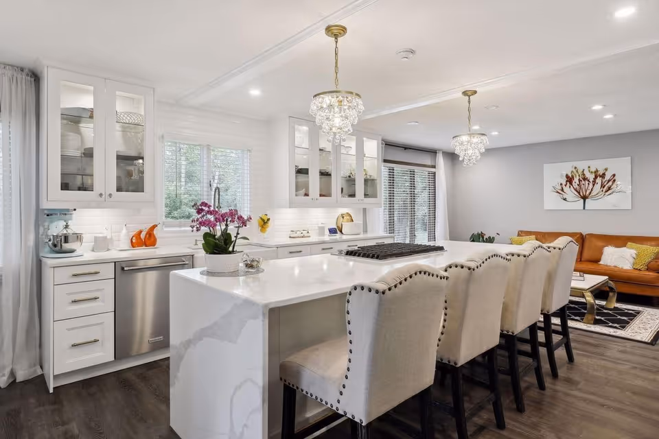 Bright, modern kitchen with island, bar stools, white cabinets, and living area in background.