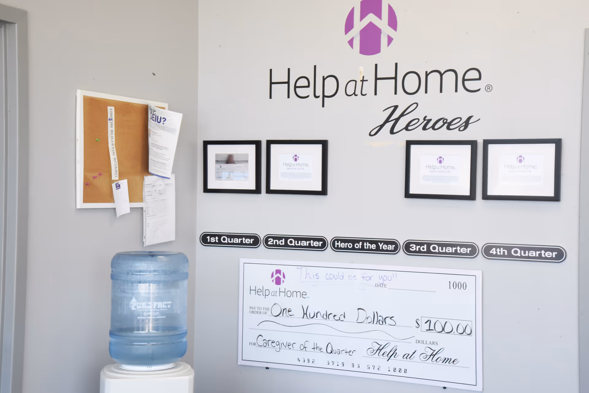Interior wall with Help at Home Heroes logo, framed certificates, bulletin board, and water cooler.