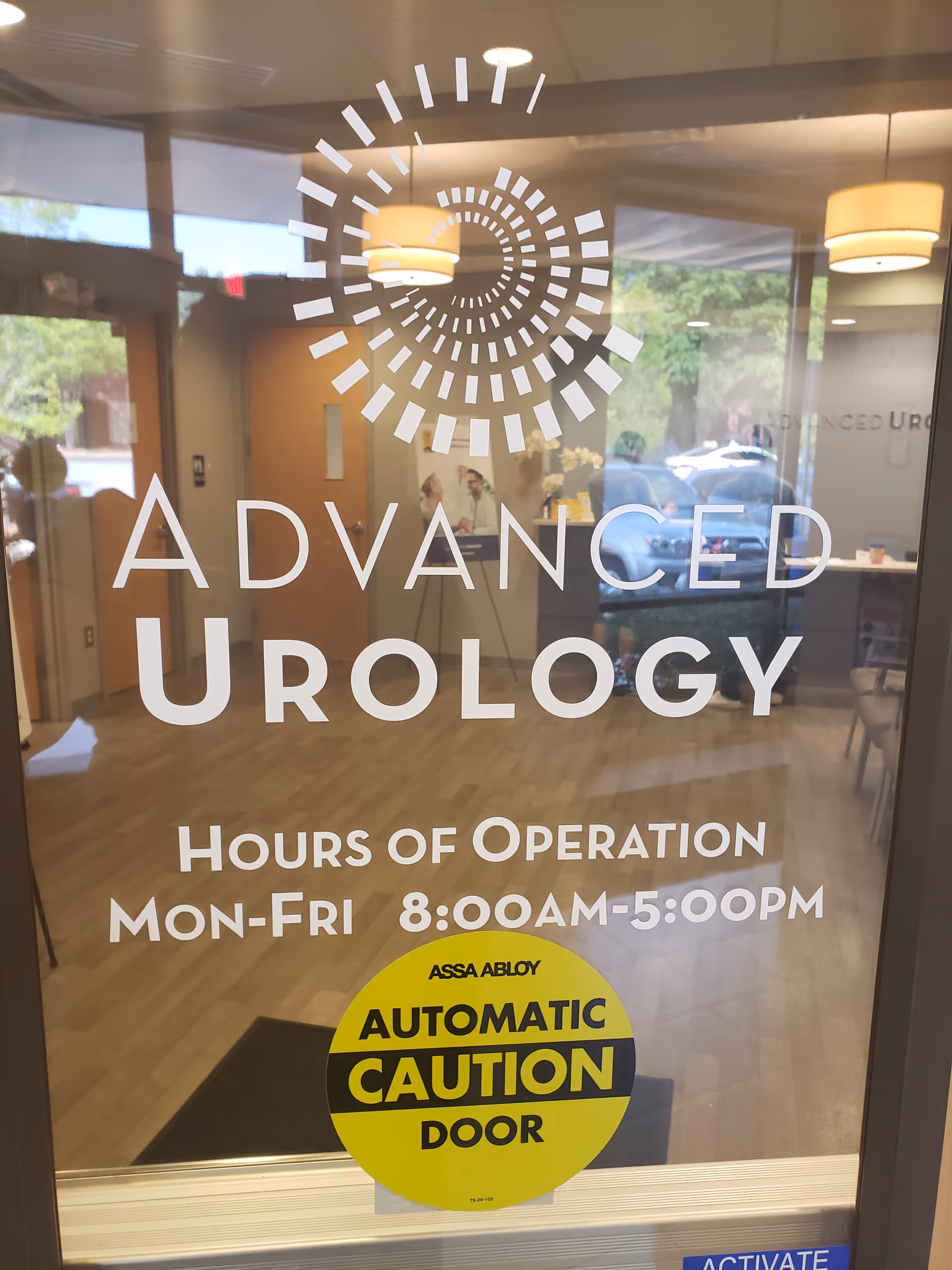 Glass door with Advanced Urology logo, hours, and yellow automatic door sticker inside lobby.