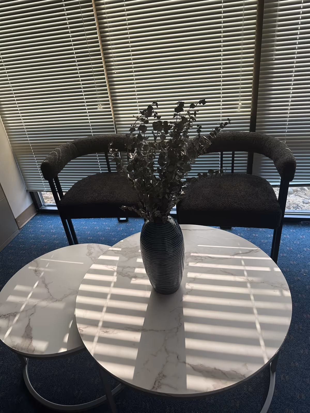 Two upholstered chairs flank a marble-round coffee table with a vase and branches.