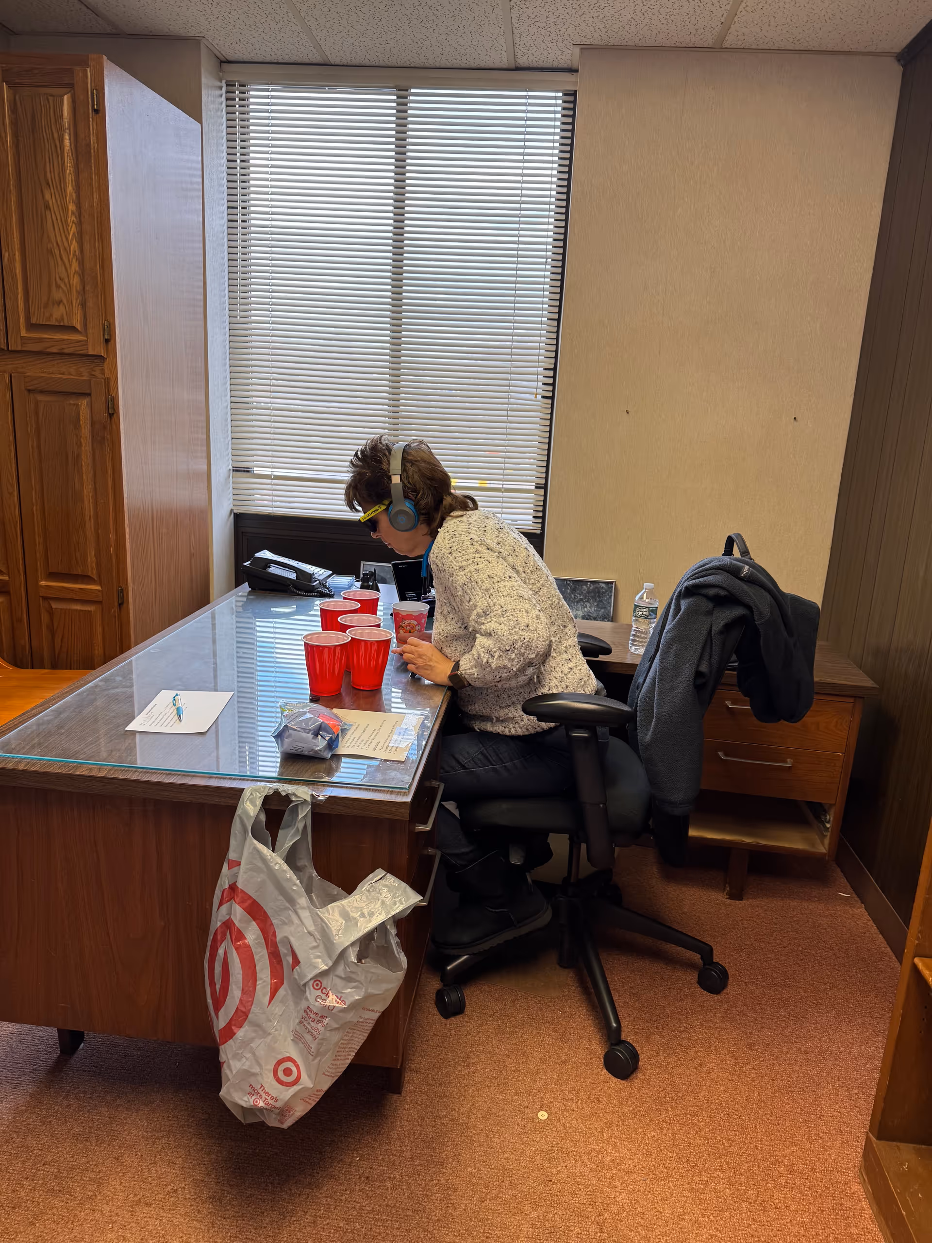 Staff member wearing headphones sits at a glass desk in a small office.