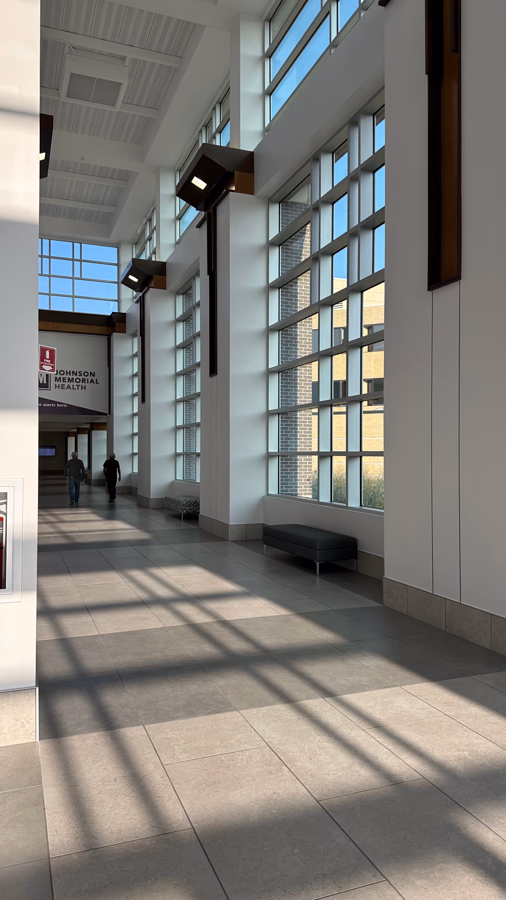 Bright modern hallway with large windows, seating benches, and two people walking toward Johnson Memorial Health.