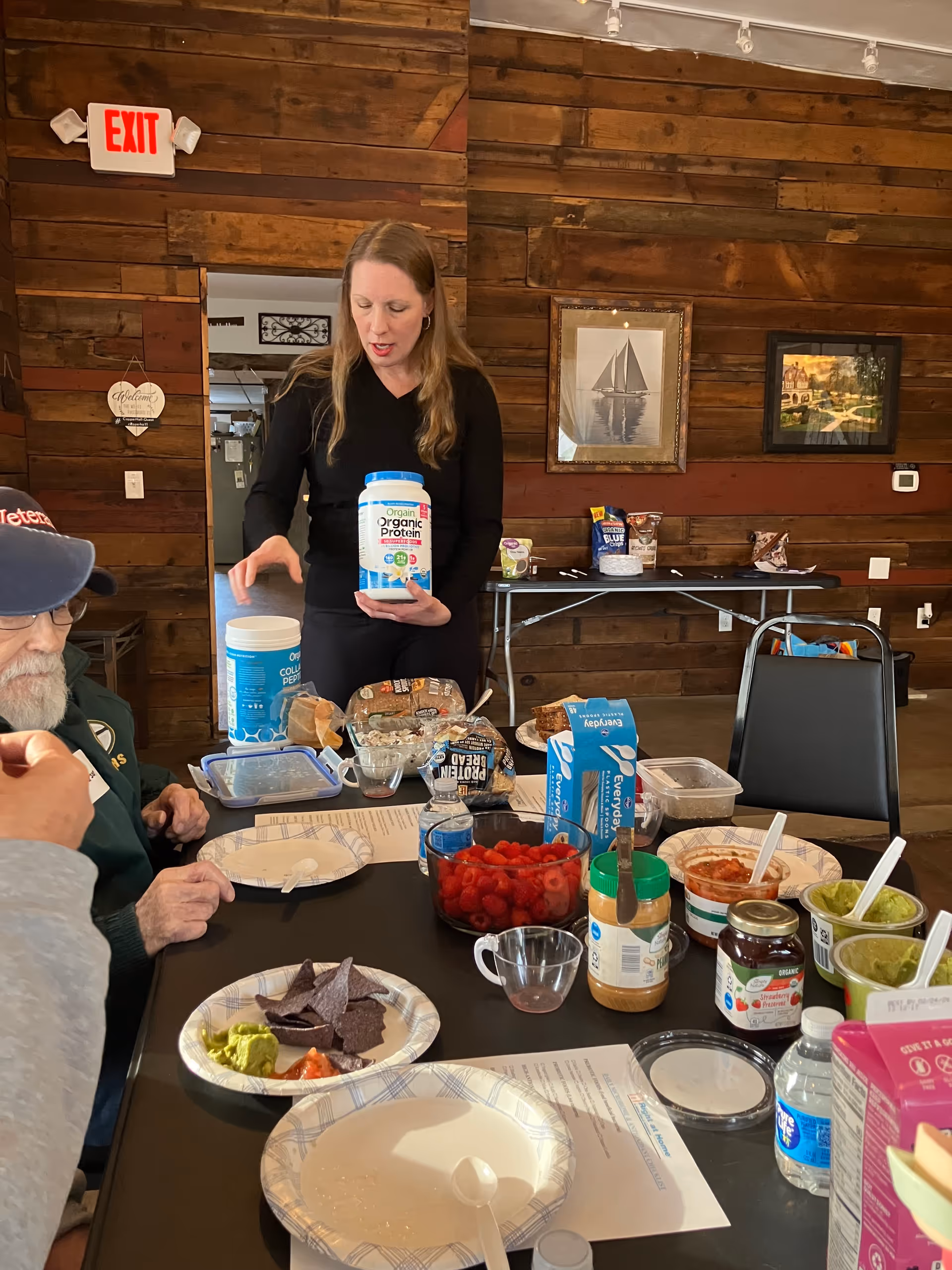 Caregiver holds a protein powder container beside a dining table with residents.