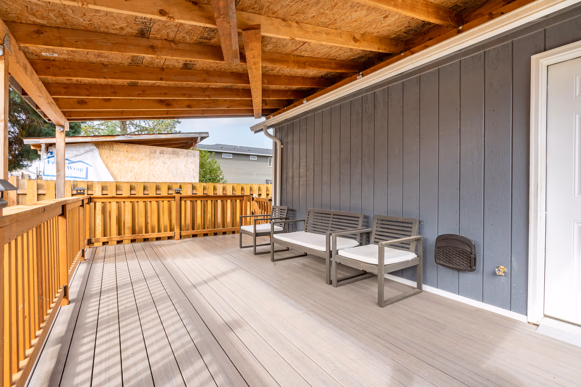 Wooden outdoor deck with blue siding, railing, and three cushioned chairs along the wall.