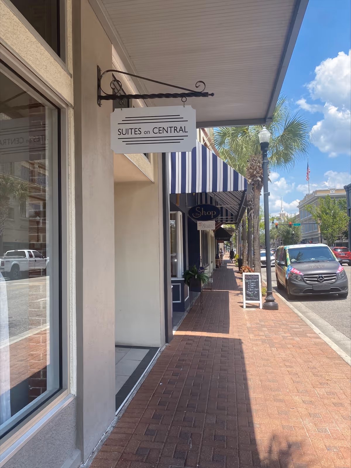 Sidewalk view of storefronts with striped awnings, a palm tree, and parked cars.