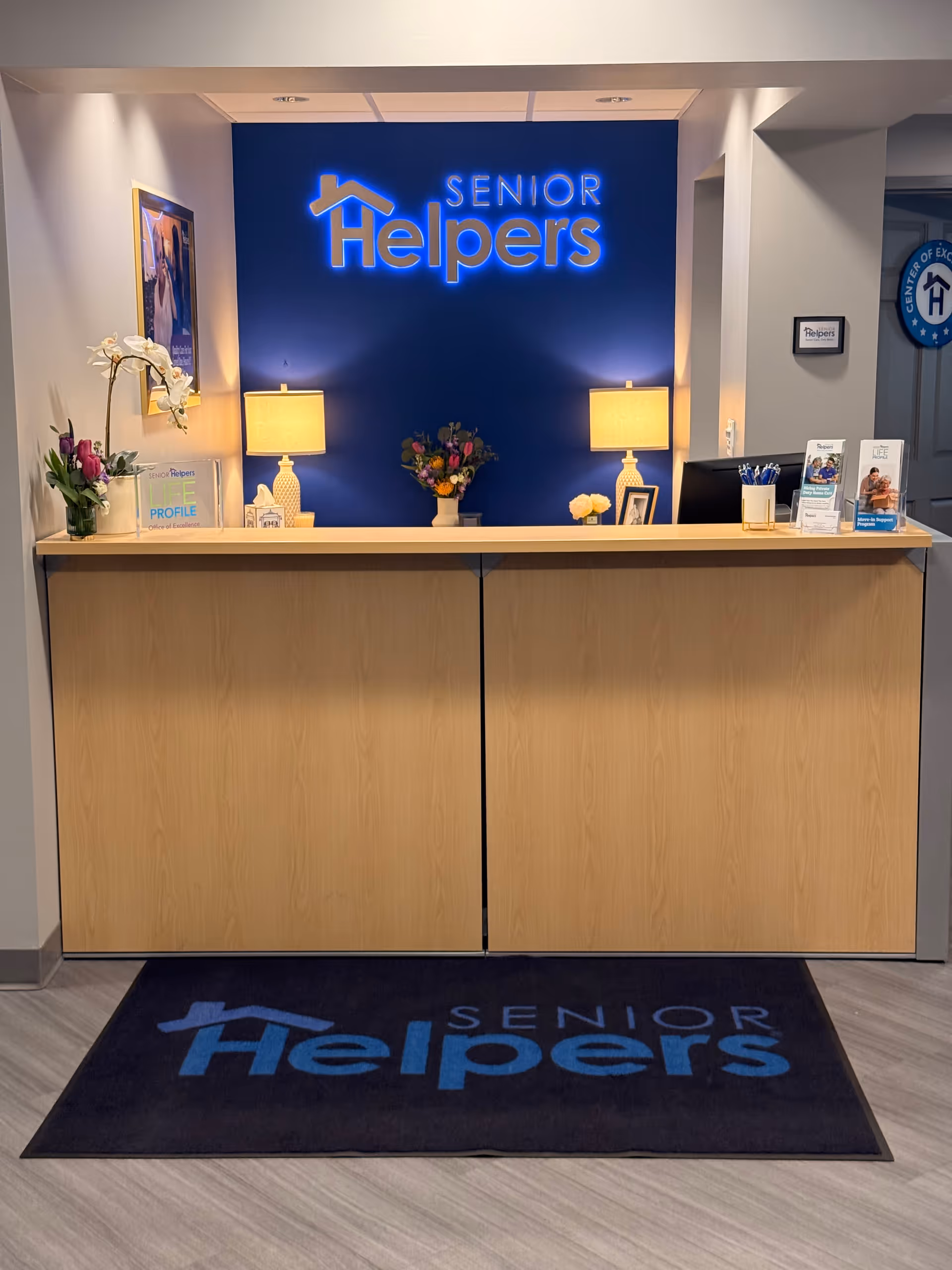Reception desk with a blue Senior Helpers sign, lamps, flowers, and brochures in a modern lobby.