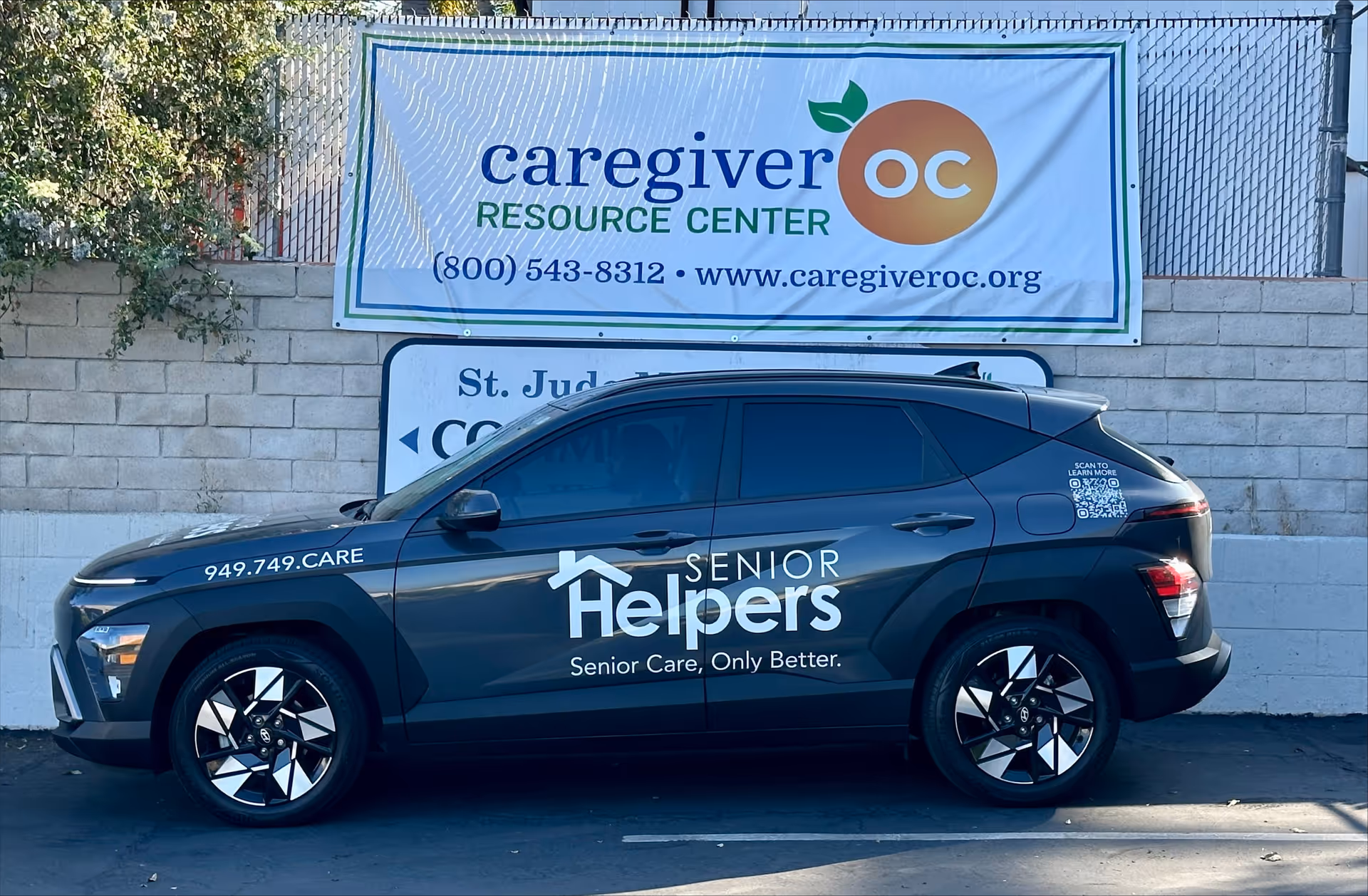 Dark gray Senior Helpers SUV parked beside a caregiver OC banner on a wall.