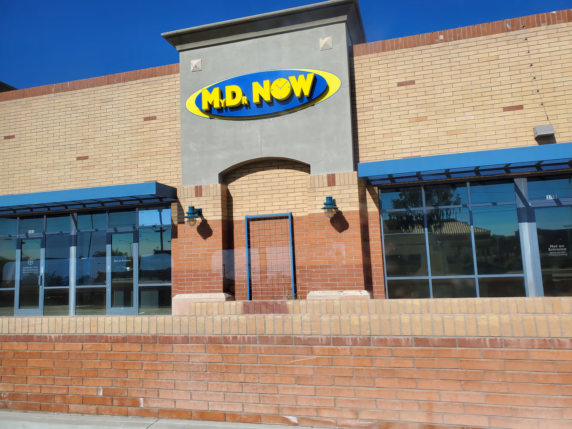 Front exterior of a brick MD NOW clinic with blue awnings and glass doors.