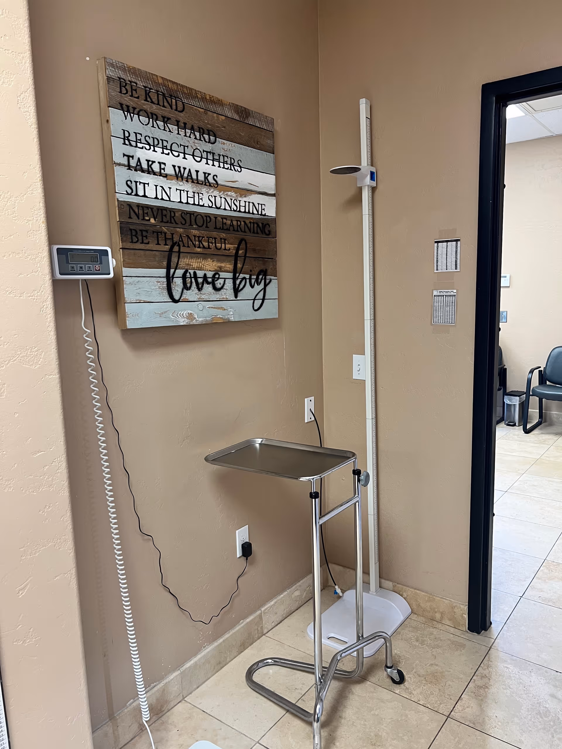 Interior hallway corner with a height scale, rolling cart, and inspirational wooden sign on beige wall.