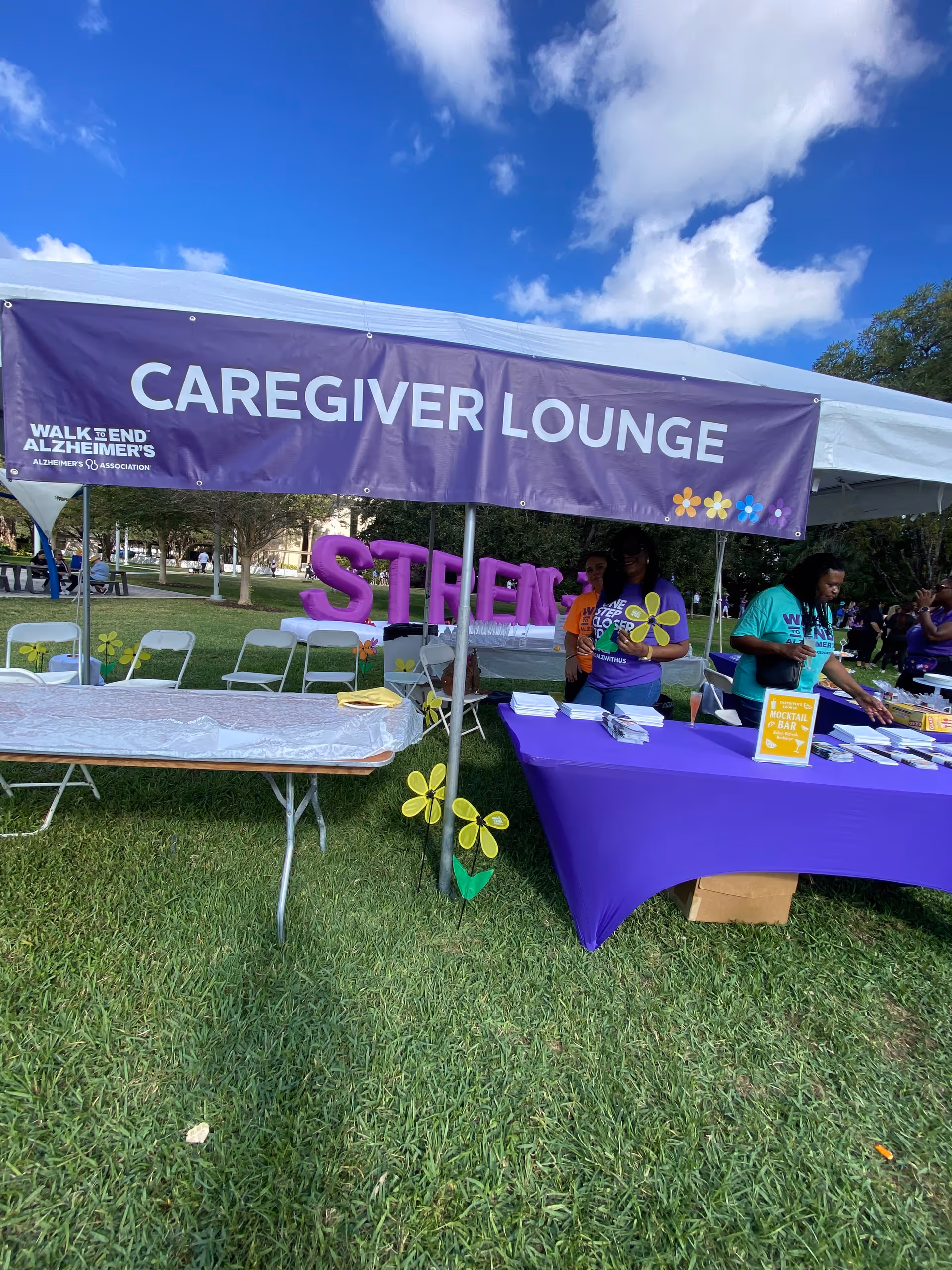 Outdoor caregiver lounge event under a tent with a purple banner and tables.