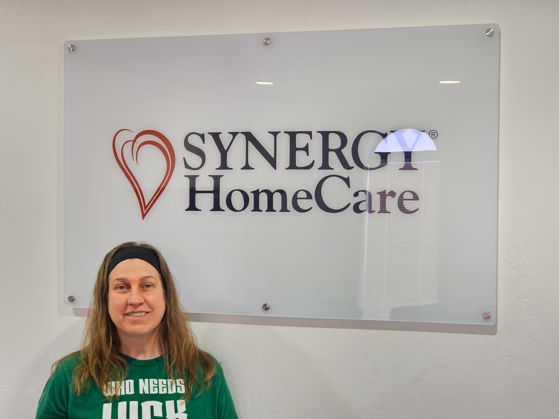 A woman stands beside a SYNERGY HomeCare sign in an interior hallway.