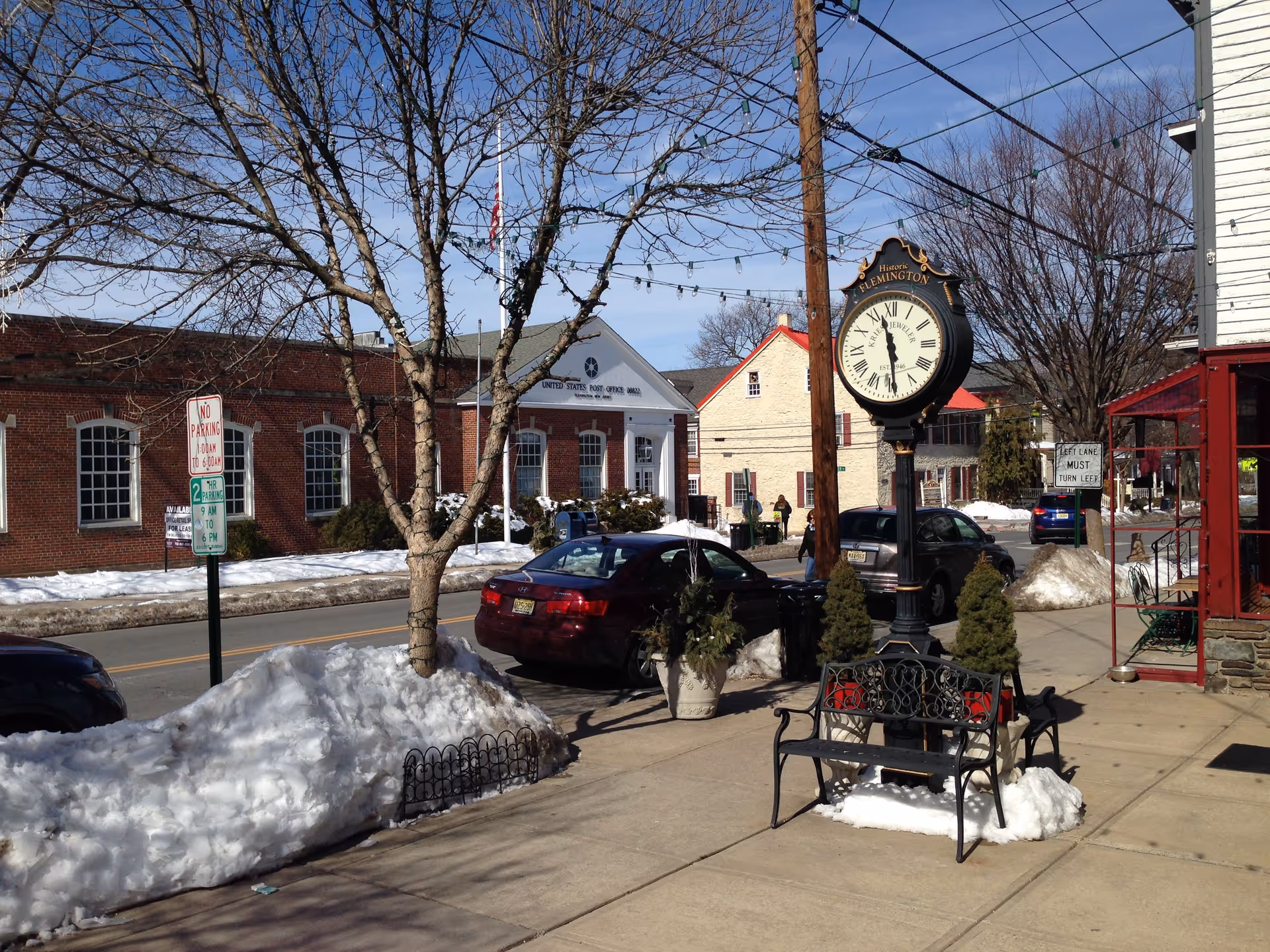 Winter street scene featuring bare trees, benches, a large clock, parked cars, and snow.