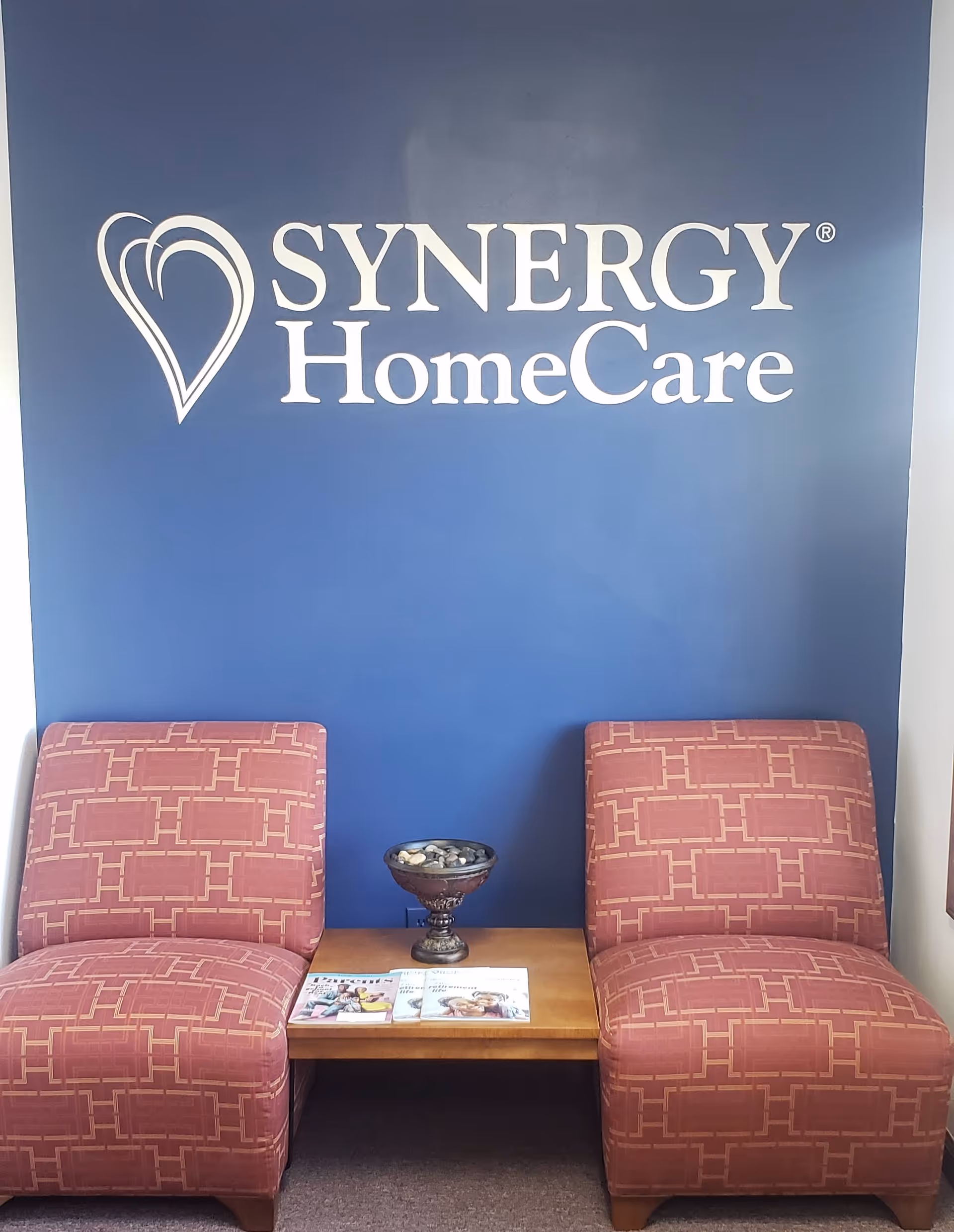 Two pink patterned chairs flank a small wooden table against a blue Synergy HomeCare wall.