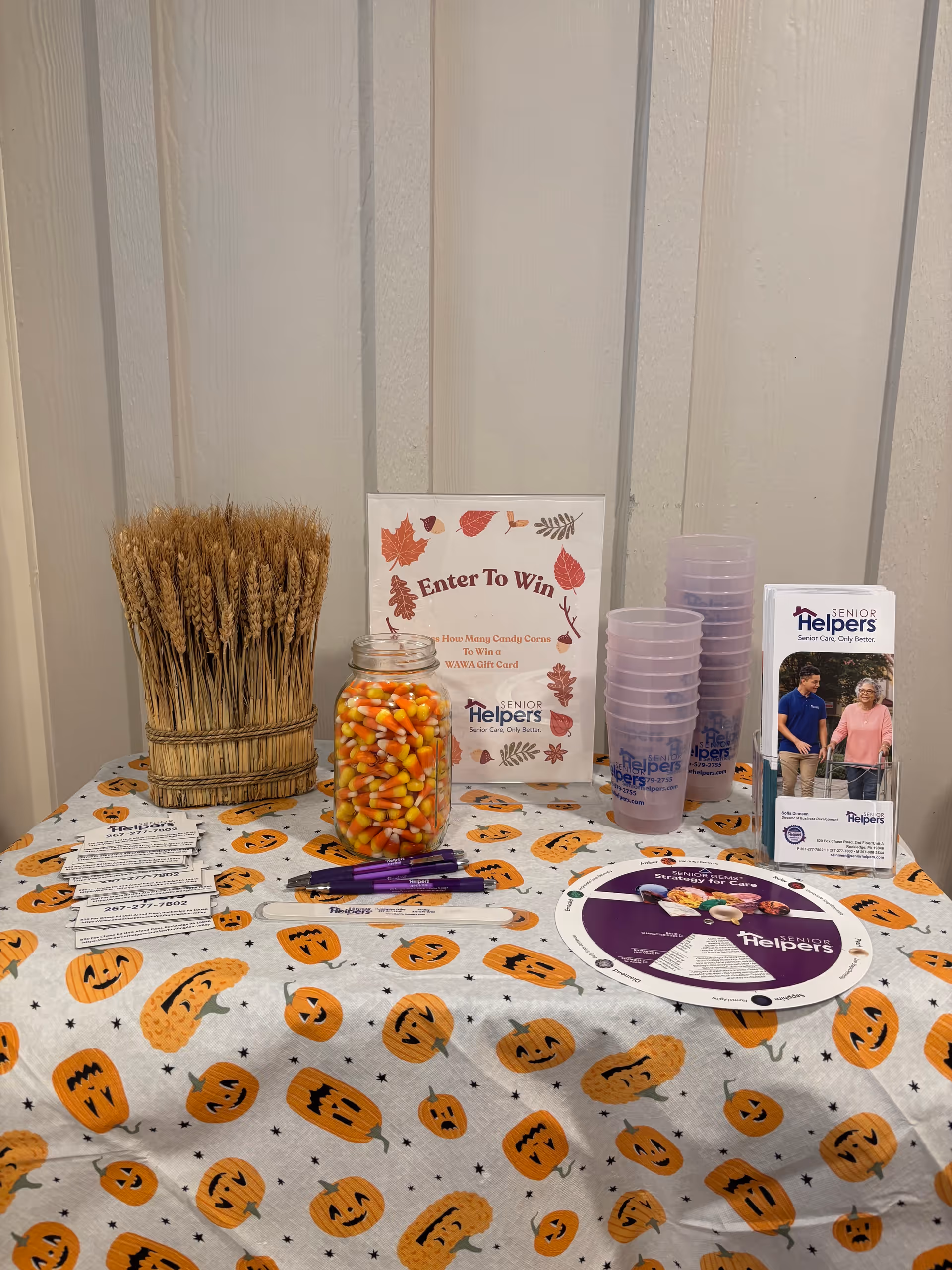 Indoor display table with Senior Helpers brochures, candy jar, pumpkin-pattern cloth, cups, and a sign.