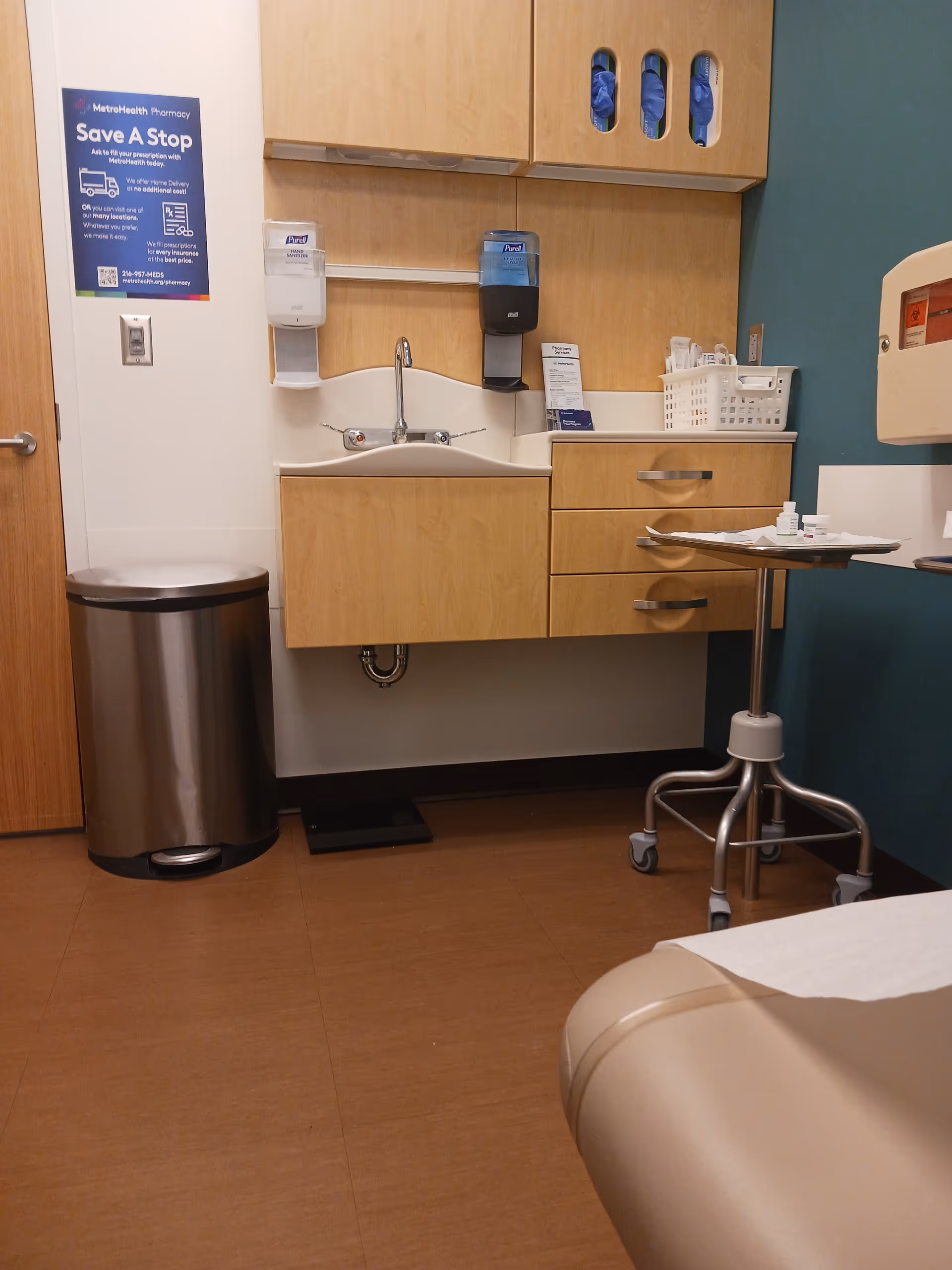 Clinic-style patient room with sink, cabinets, dispensers, a rolling table, and a bed.