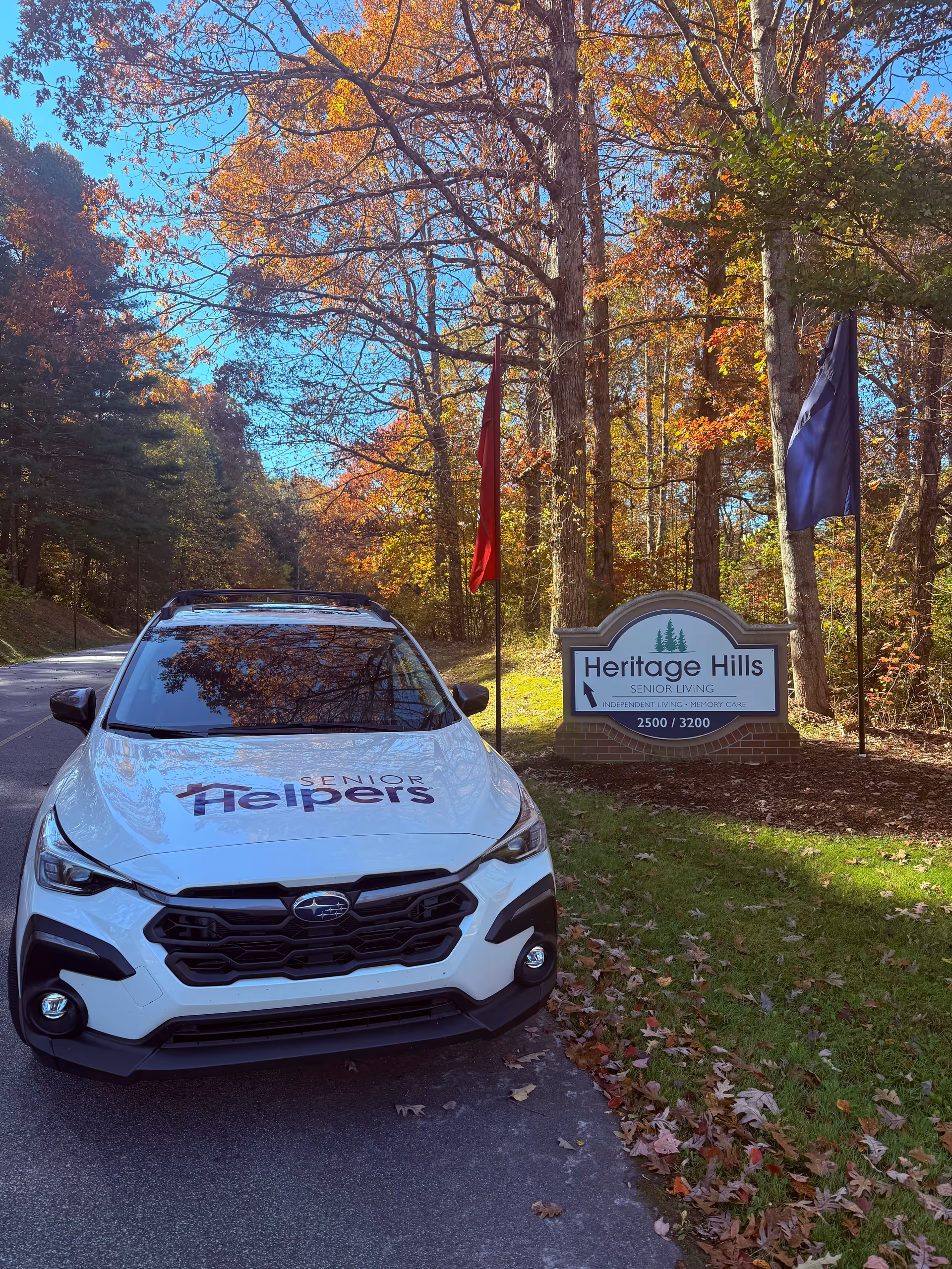 A white Senior Helpers vehicle parked beside a Heritage Hills Senior Living sign among fall trees.