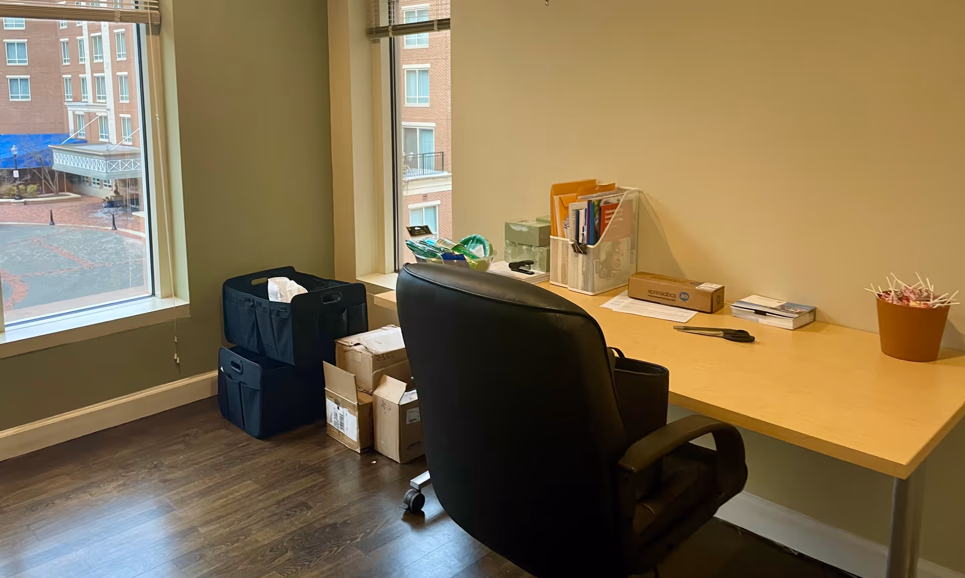 Office workspace with a desk, rolling chair, files, and supplies by a window.