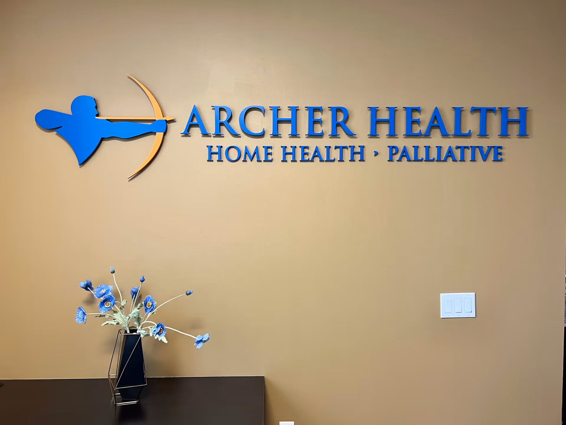 Interior wall signage reads Archer Health: Home Health and Palliative, with a blue flower vase.