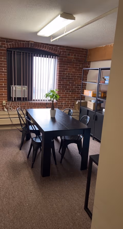 Brick walled room with a dark dining table and metal chairs, potted plant centerpiece.