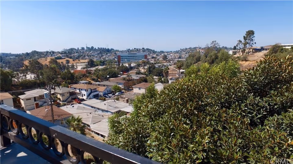 Balcony railing and lush bushes overlook a hillside residential area with rooftops and trees.