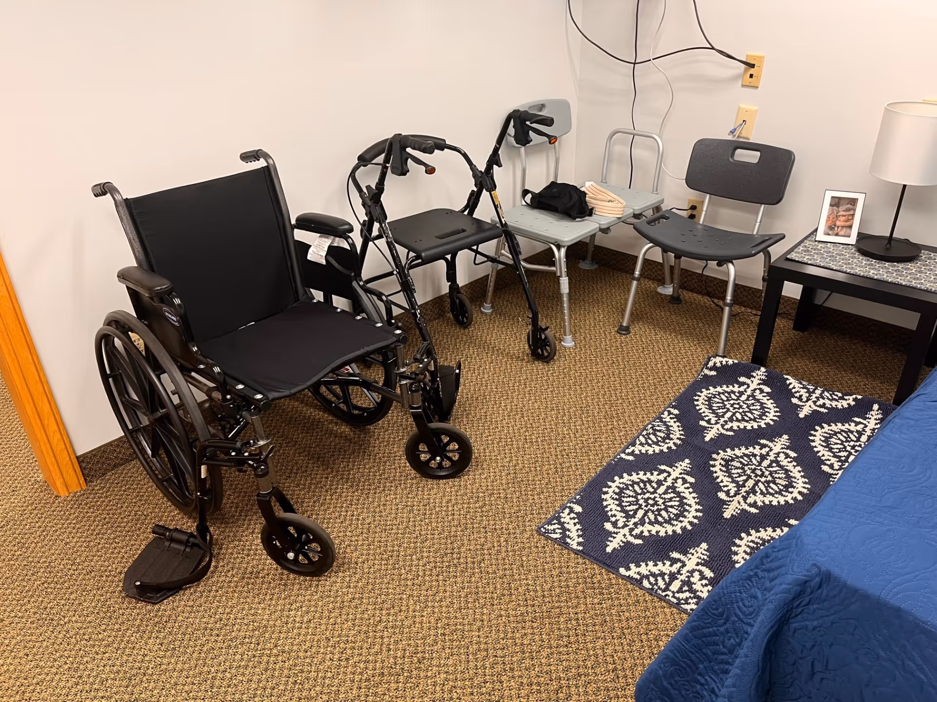 Interior senior living bedroom with wheelchair, walker, chairs, bed, and nightstand.
