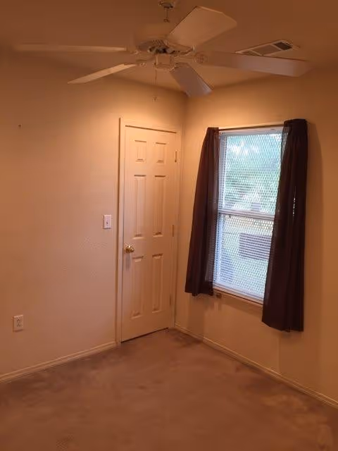 Empty beige room with ceiling fan, door, window with dark curtains, and carpet.