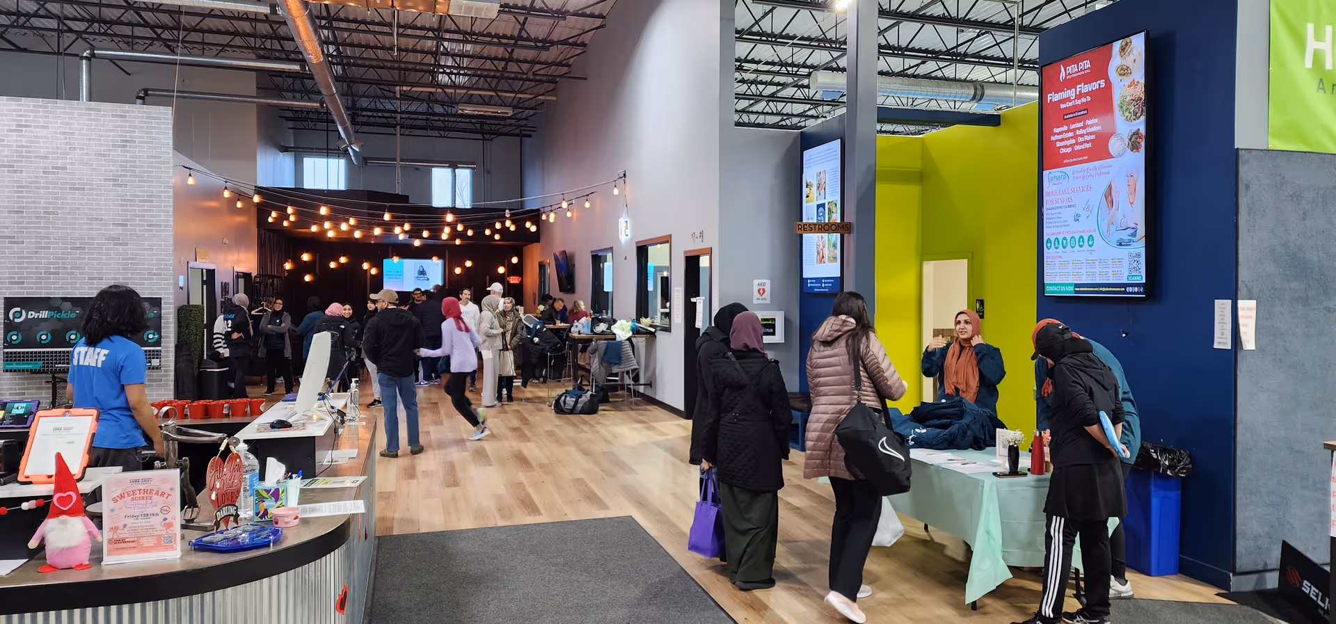 Busy indoor community area with staff, visitors, tables, screens, and warm string lights.