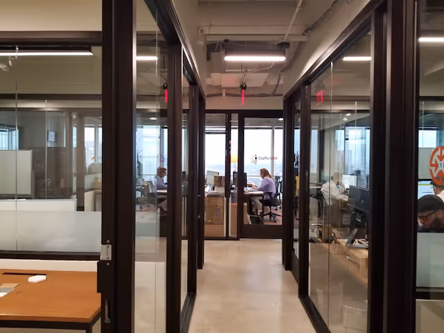 Glass-walled office hallway with people seated at desks in a bright interior.