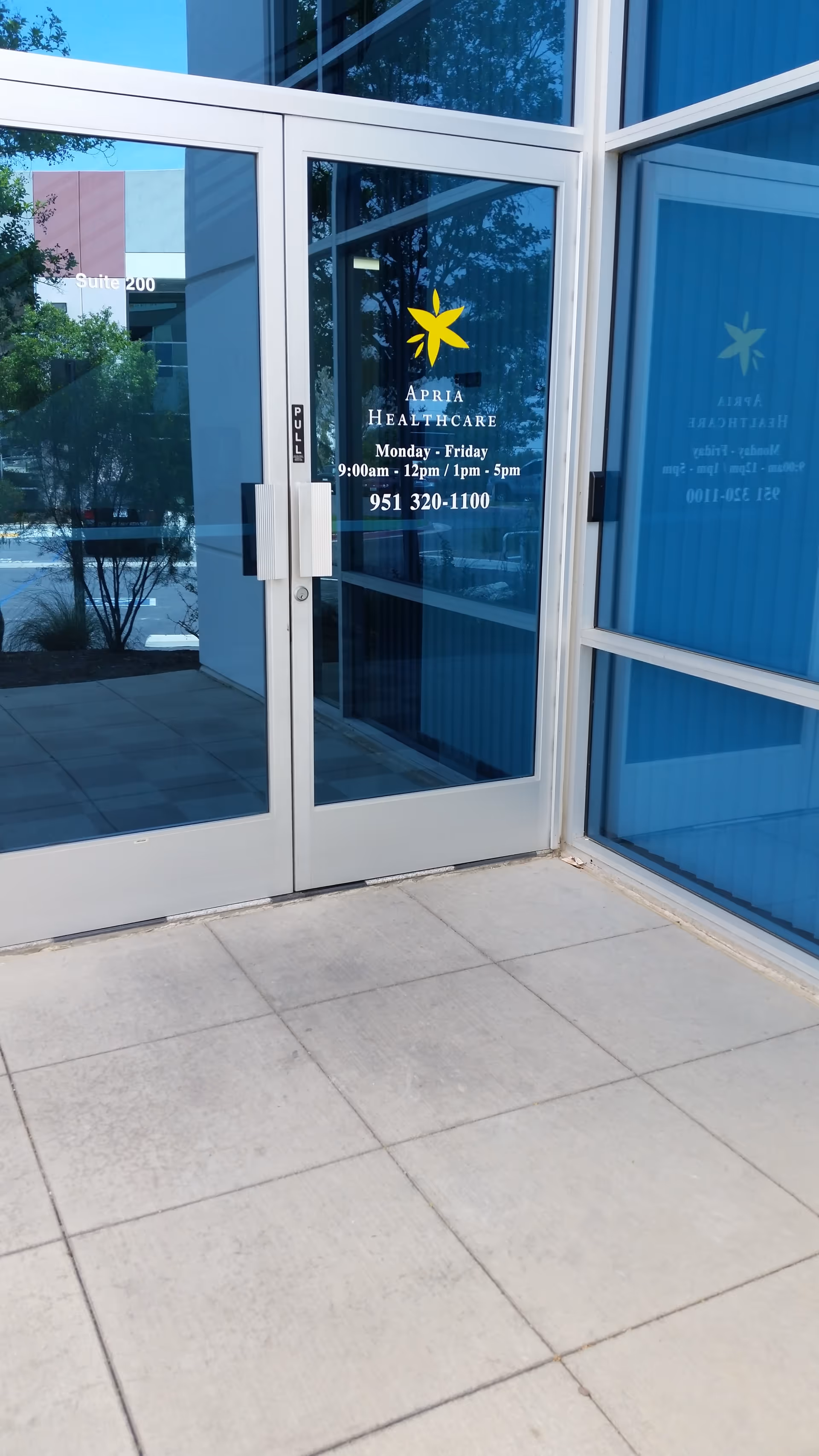 Glass entrance door with Apria Healthcare logo and hours visible on building front.