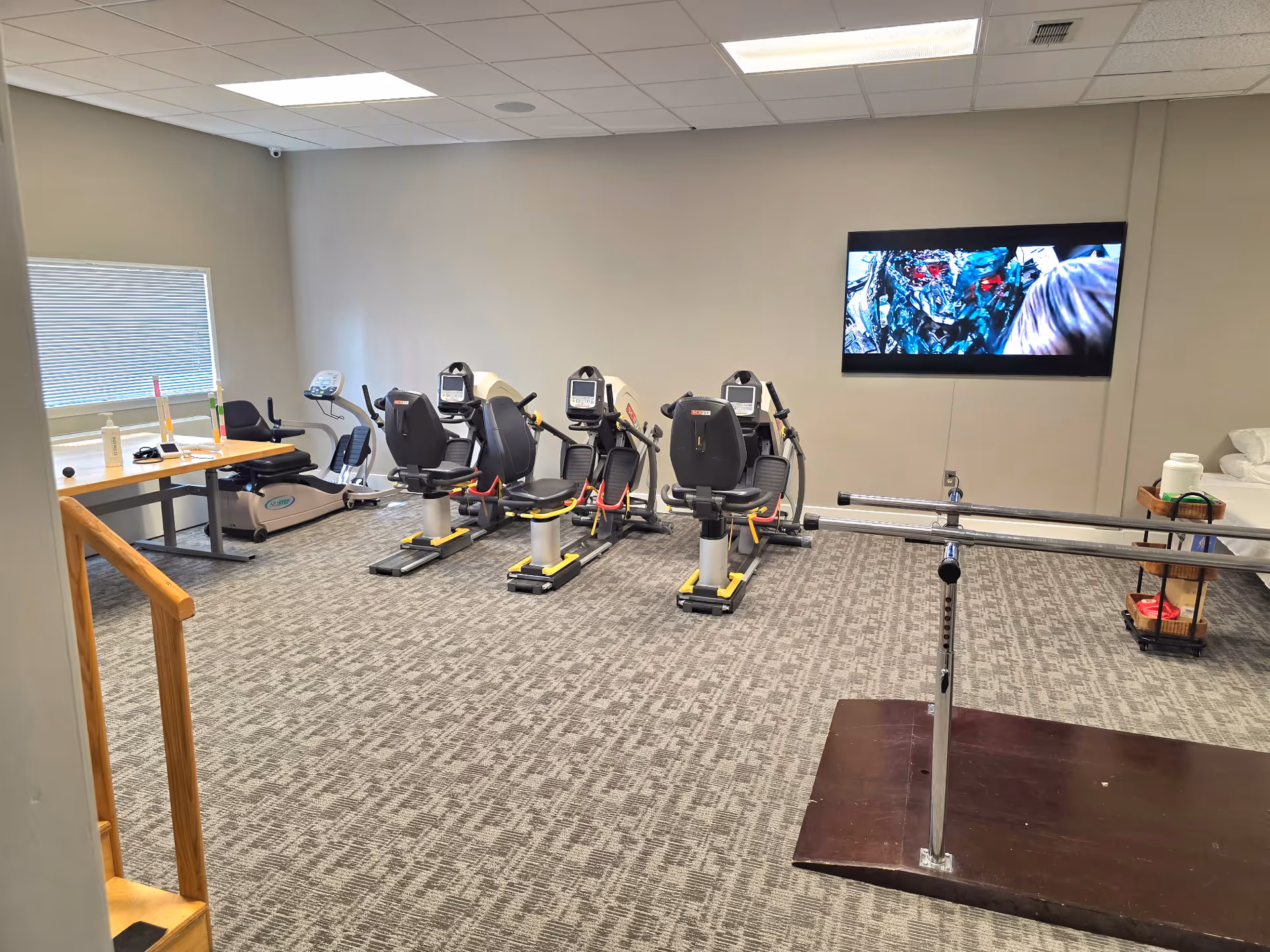 Empty senior exercise room with stationary bikes, a TV, and a railing.