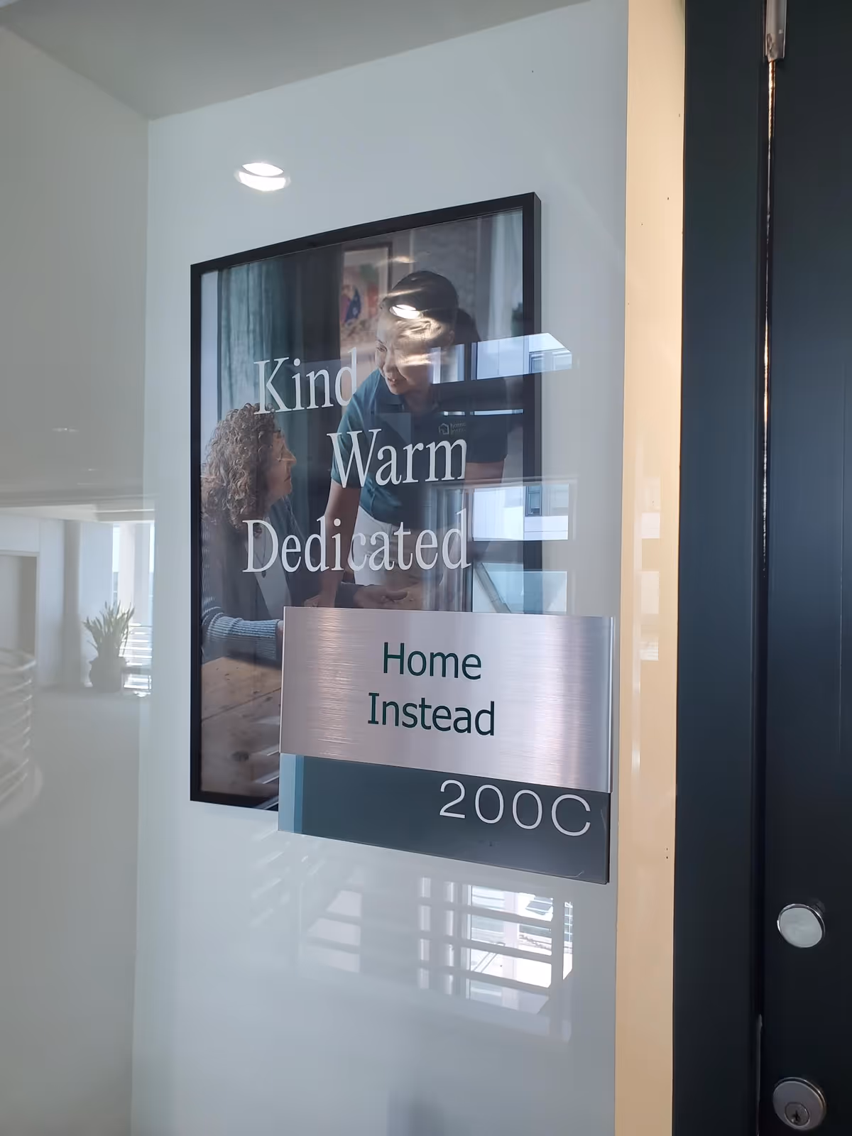 Glass door with a Home Instead sign and poster reading Kind Warm Dedicated