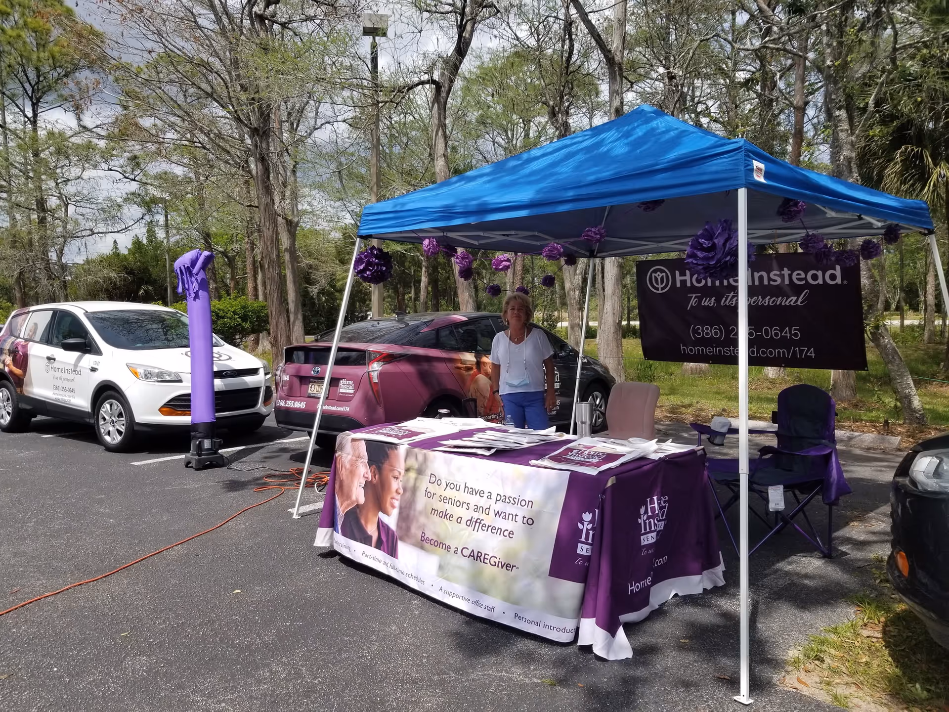 Outdoor Home Instead information booth under a blue canopy with a table and a staff member.