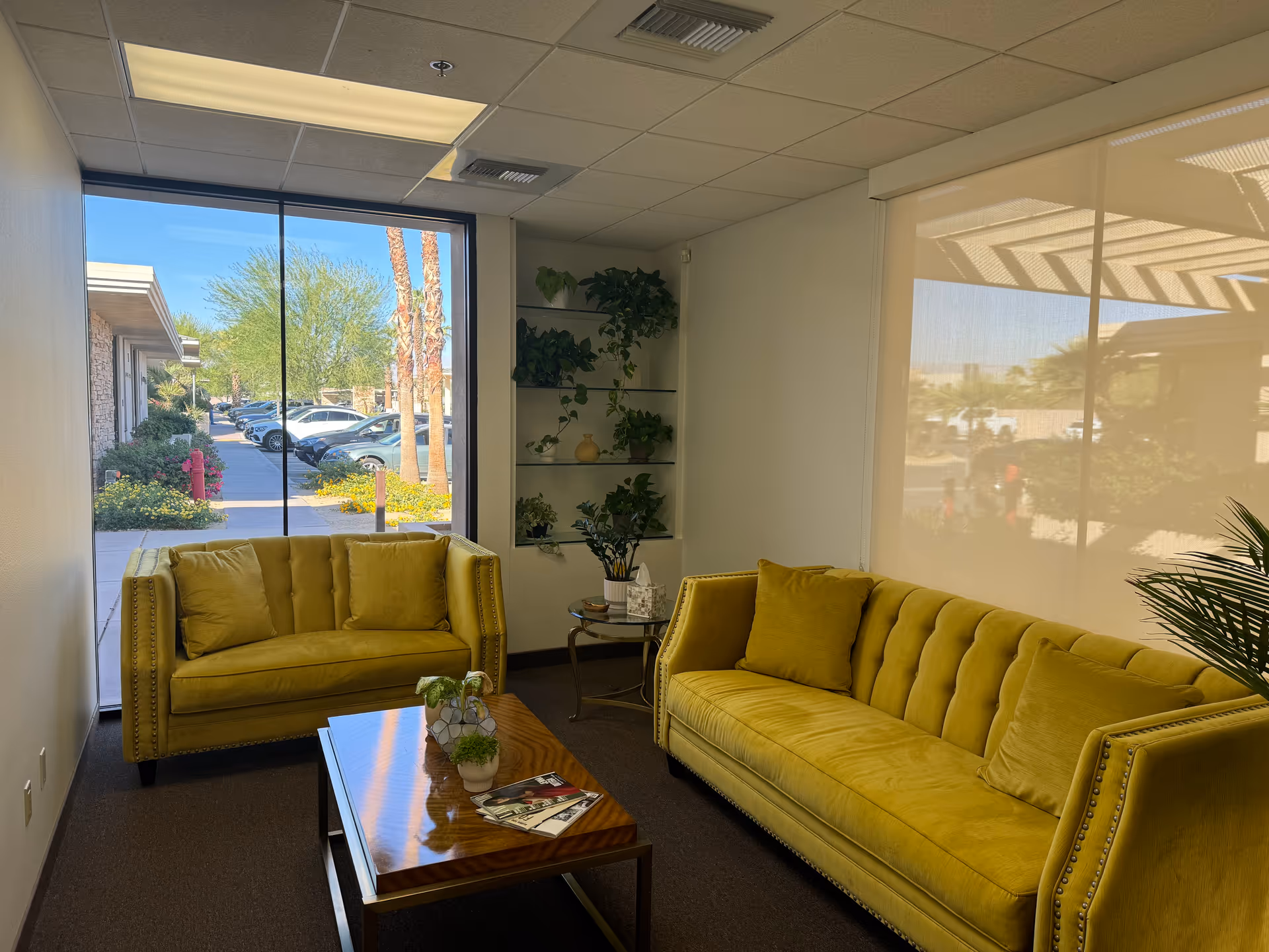 Bright lounge area with two yellow sofas, coffee table, plants, and large window view.