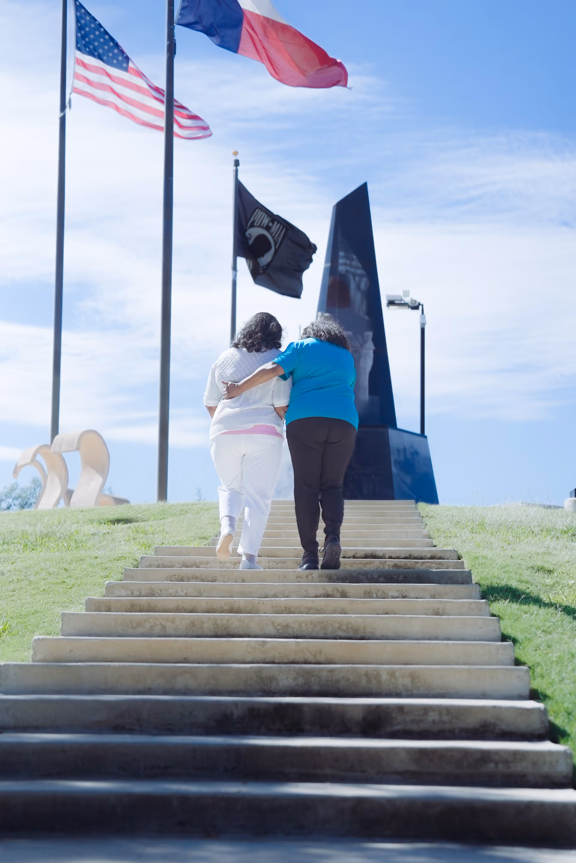 Two people walk up stone steps toward a memorial sculpture with flags in an outdoor area.