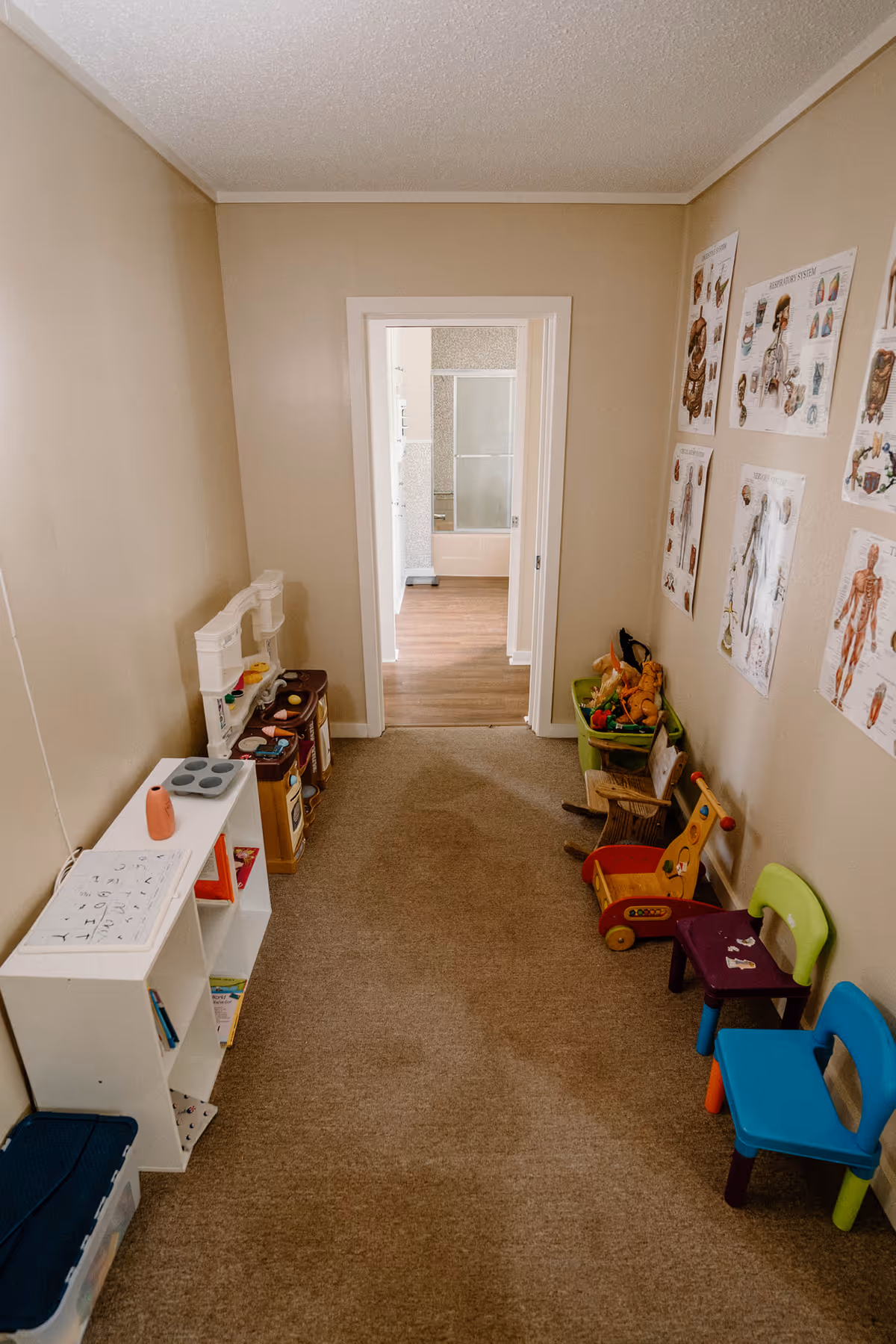 Narrow beige hallway with children's play area, toys, posters, and small chairs along wall.