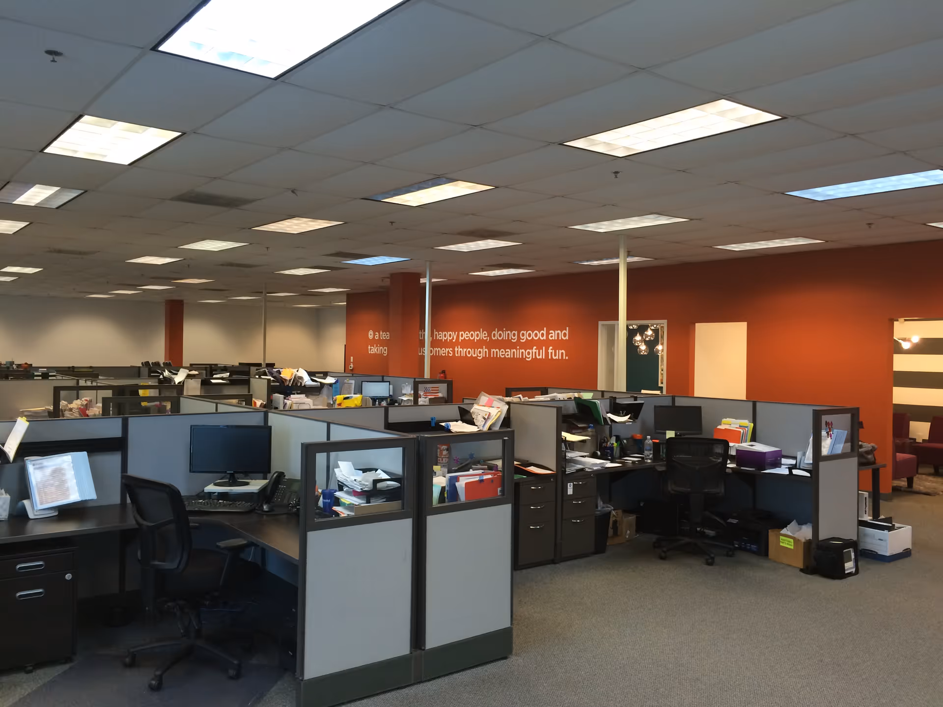 Open-plan office with cubicles, desks, computers, and an orange accent wall.