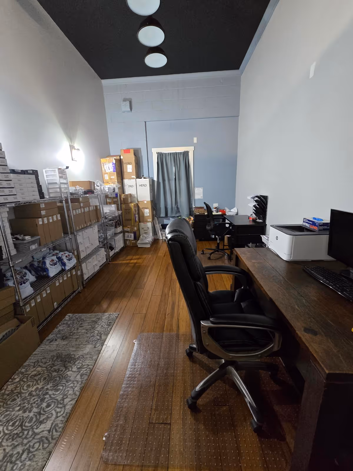 Office workspace in a storage area with desks, chairs, shelves, and boxes.