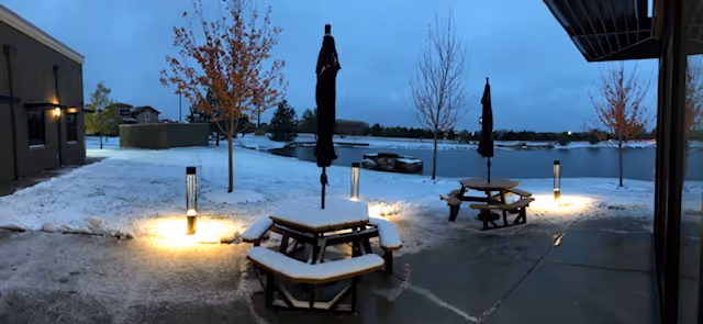 Snowy outdoor patio with picnic tables, closed umbrellas, and a lakeside view.