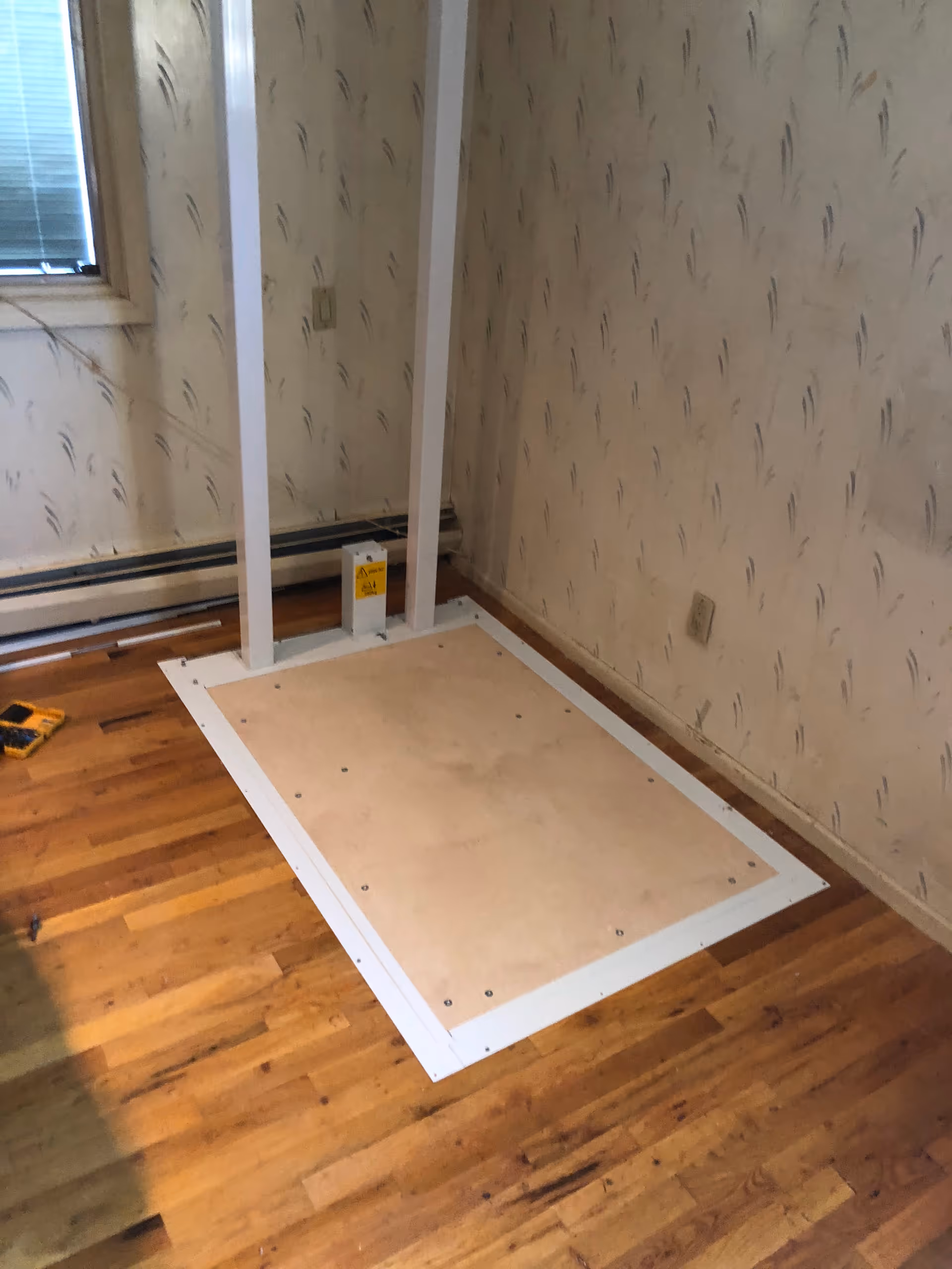 Interior room with wallpaper, hardwood floor, a large square panel on the floor and white posts.