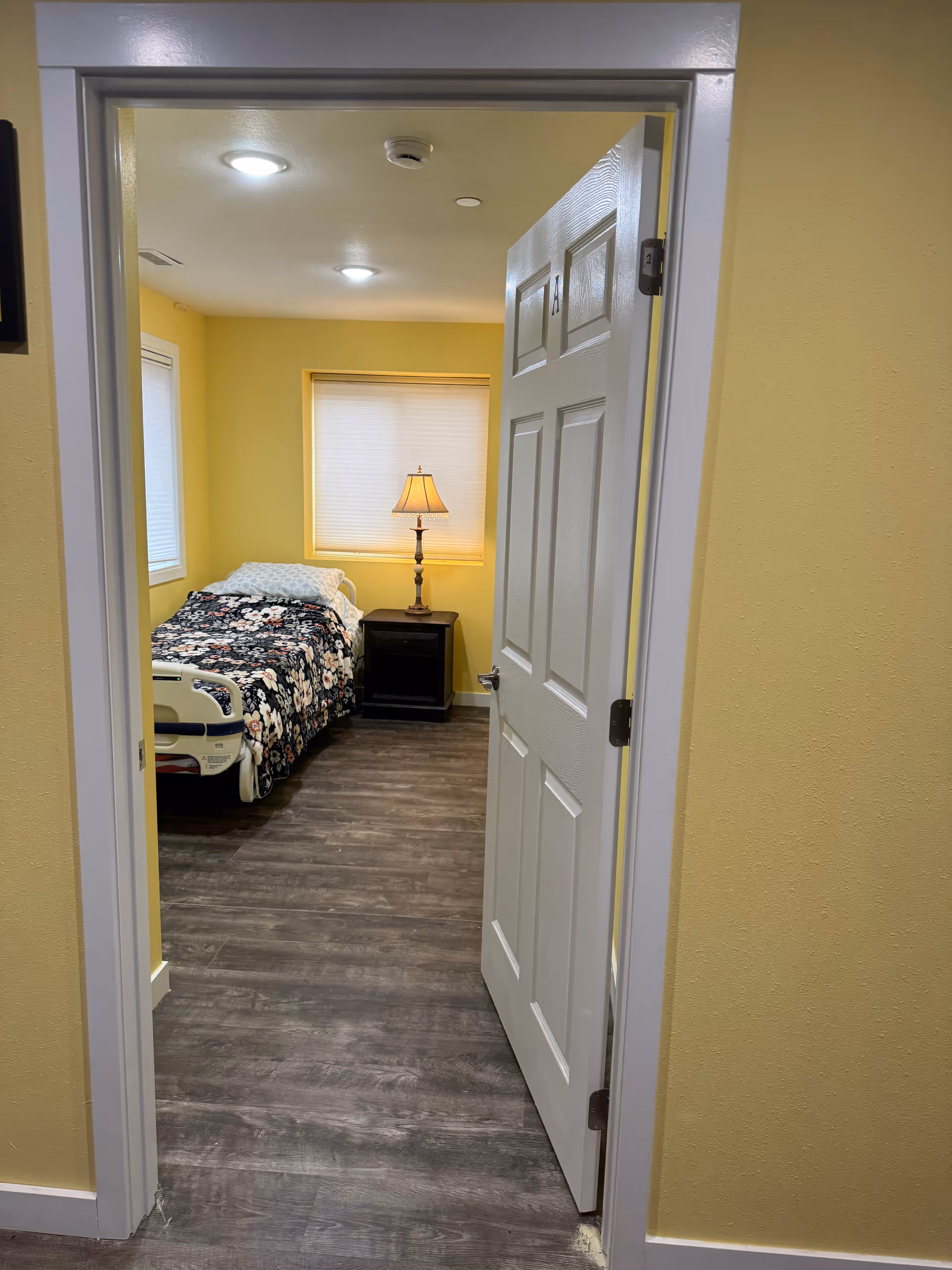 Yellow-walled bedroom with floral bedspread, nightstand, lamp, and window visible through an open door.