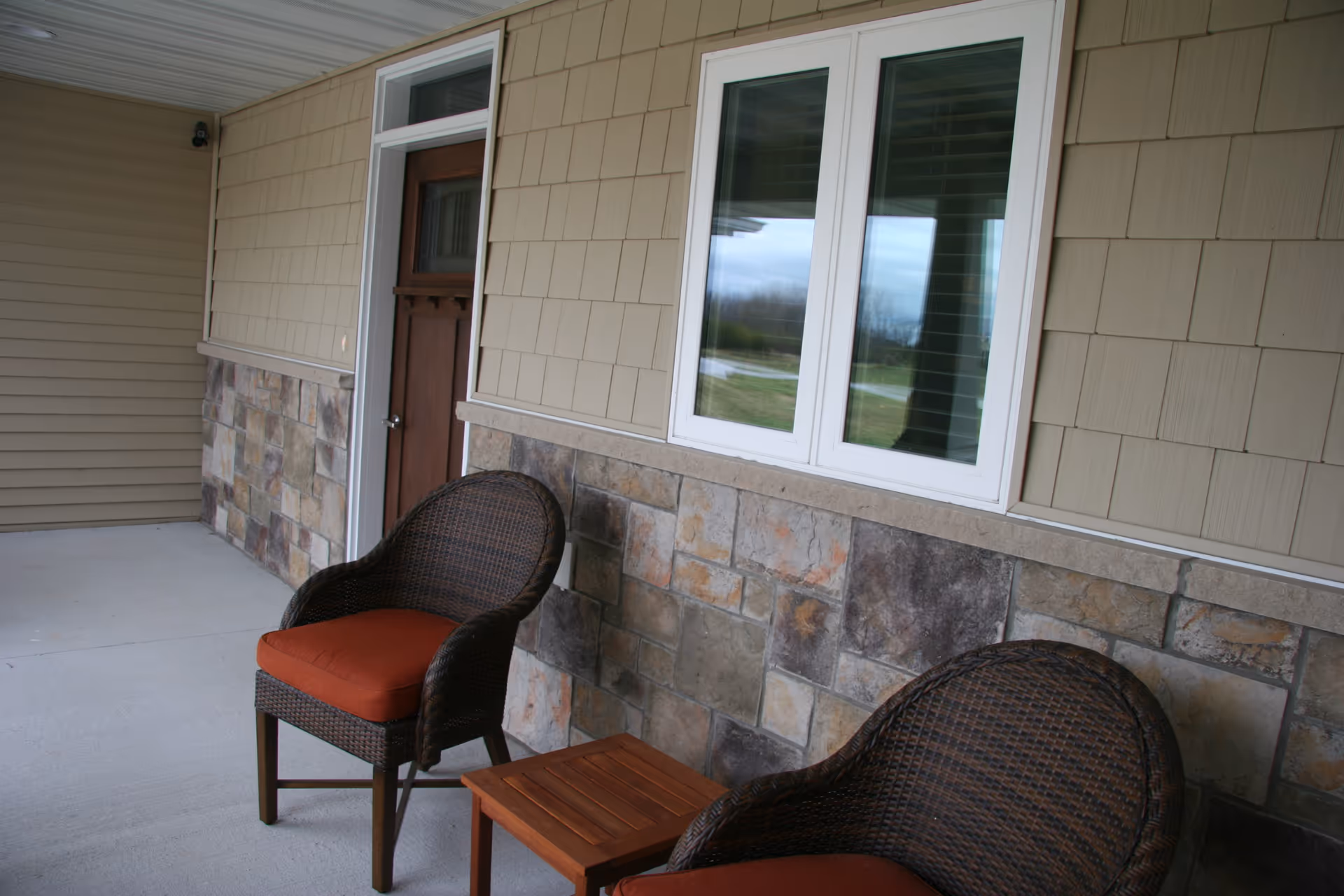 Outdoor porch with wicker chairs and a small table against beige siding and stone wall.