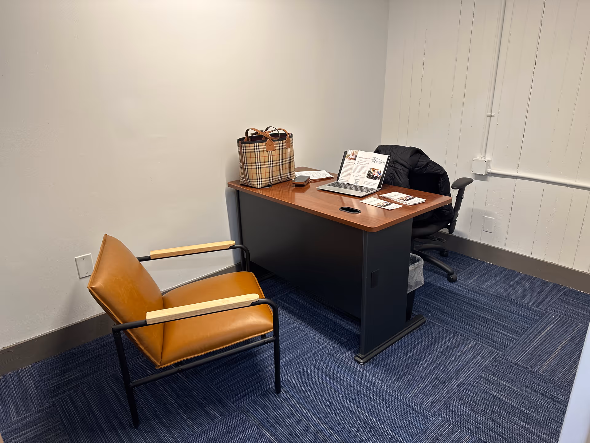 Office desk with laptop, brochures, and a bag beside a brown chair in a small room.