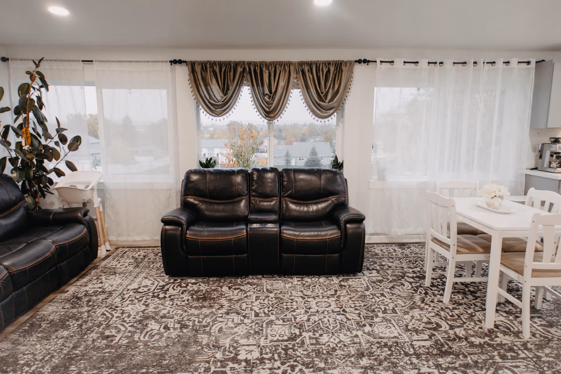 A cozy living room with black leather seating, white dining table, and sheer curtains.